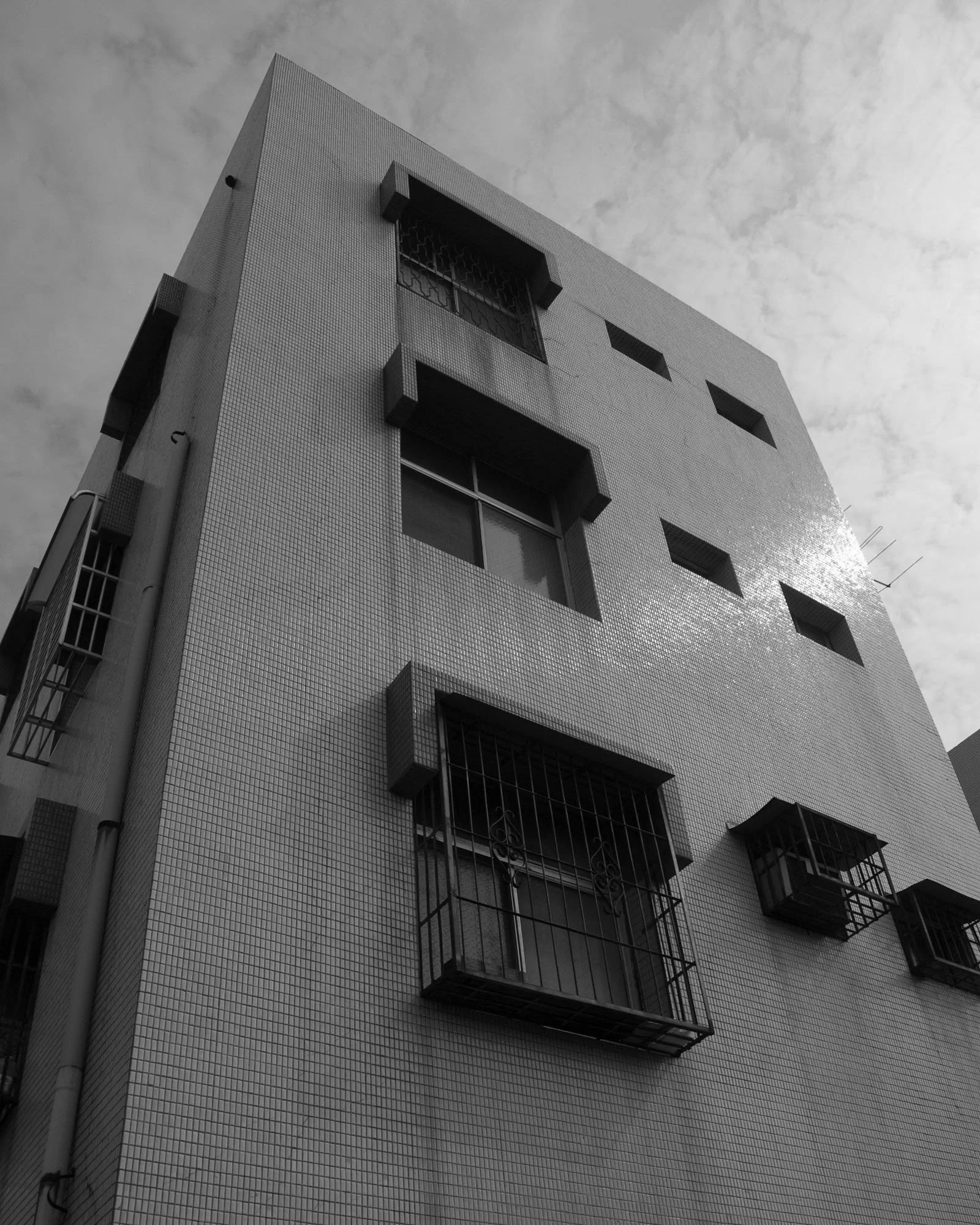 Black and white photo of a tall building with small, barred balconies and multiple windows, under a cloudy sky.