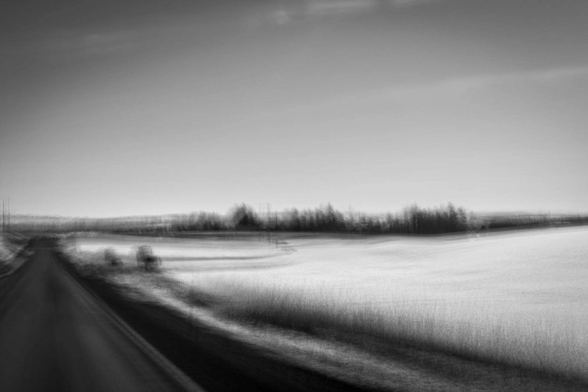 A black and white image of a countryside road with fields on either side, blurred trees in the distance, and a cloudy sky.