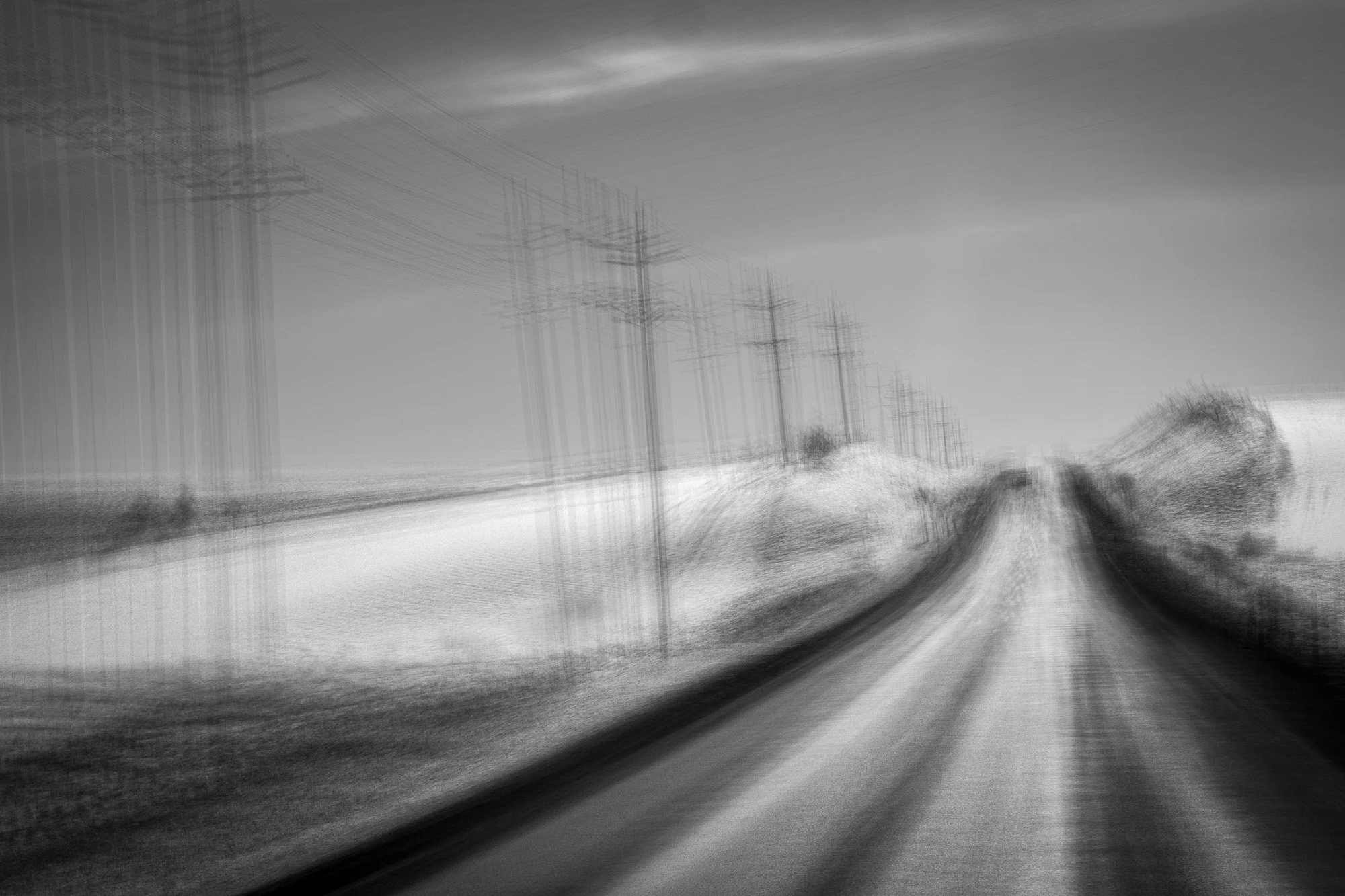 Black and white photo of a rural road with power lines and poles on either side, seen from a moving vehicle creating a blurred, streaked effect.