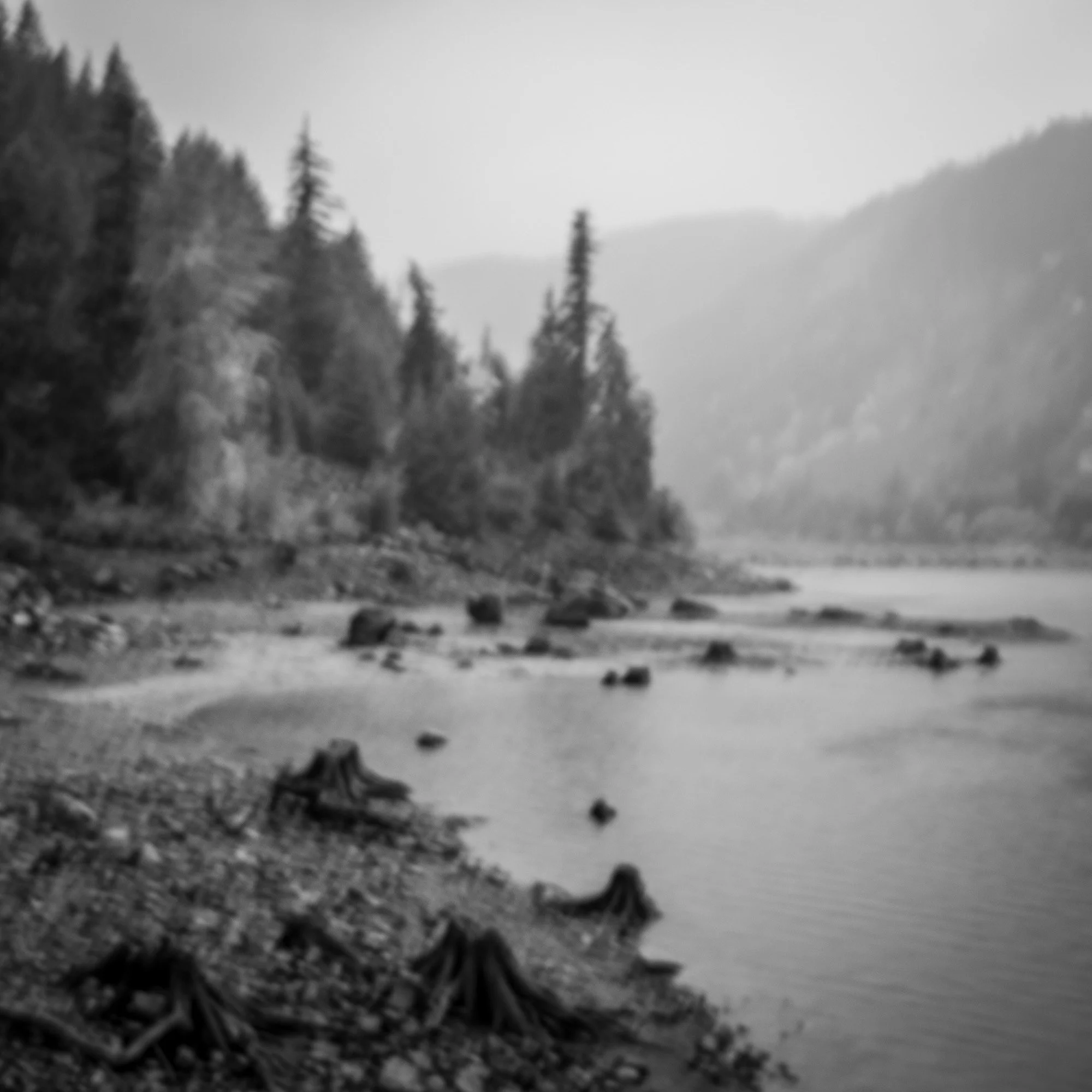 Black and white photo of a river flowing through a forested landscape with mountains in the background.