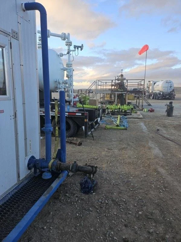 Oil or fuel transport truck on a dirt airfield with equipment and a person working on a platform in the background, cloudy sky above.