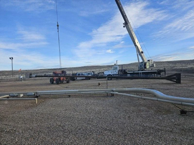 Construction site with a large crane, vehicles, and a pipeline in foreground, under a partly cloudy sky.