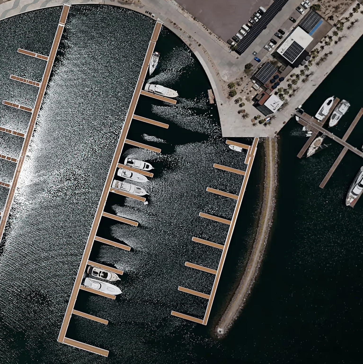 Aerial view of a marina with boats docked at floating piers, adjacent to a parking lot and waterfront walkway.