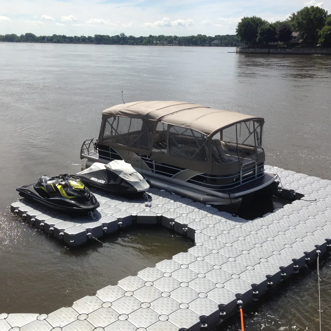 A boat docked on a floating pier with a jet ski and a personal watercraft on the dock, with a river and trees in the background.