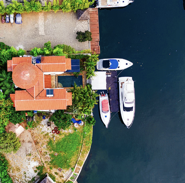 Aerial view of a house next to a waterway with two boats docked at the dock, surrounded by lush green trees and a concrete parking lot.