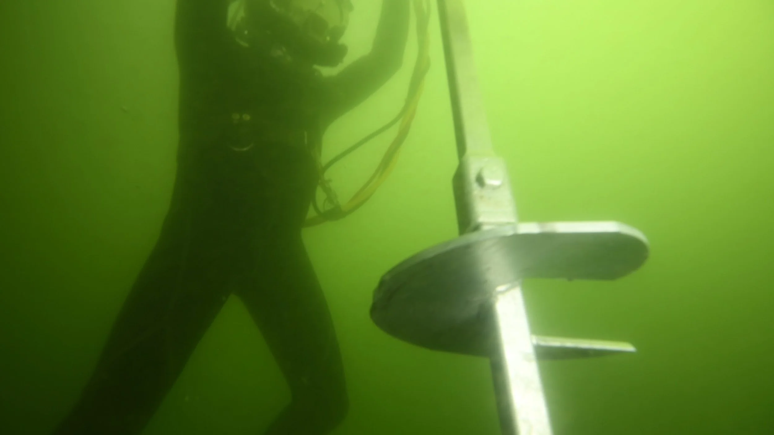 Underwater scene with a person in a wetsuit using an underwater trawl or net against a yellow-green background.