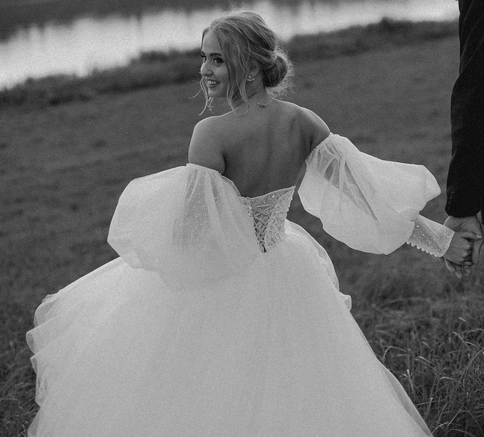 A black-and-white photo of a smiling woman in a wedding dress, holding hands with a man outdoors on a grassy area near water.