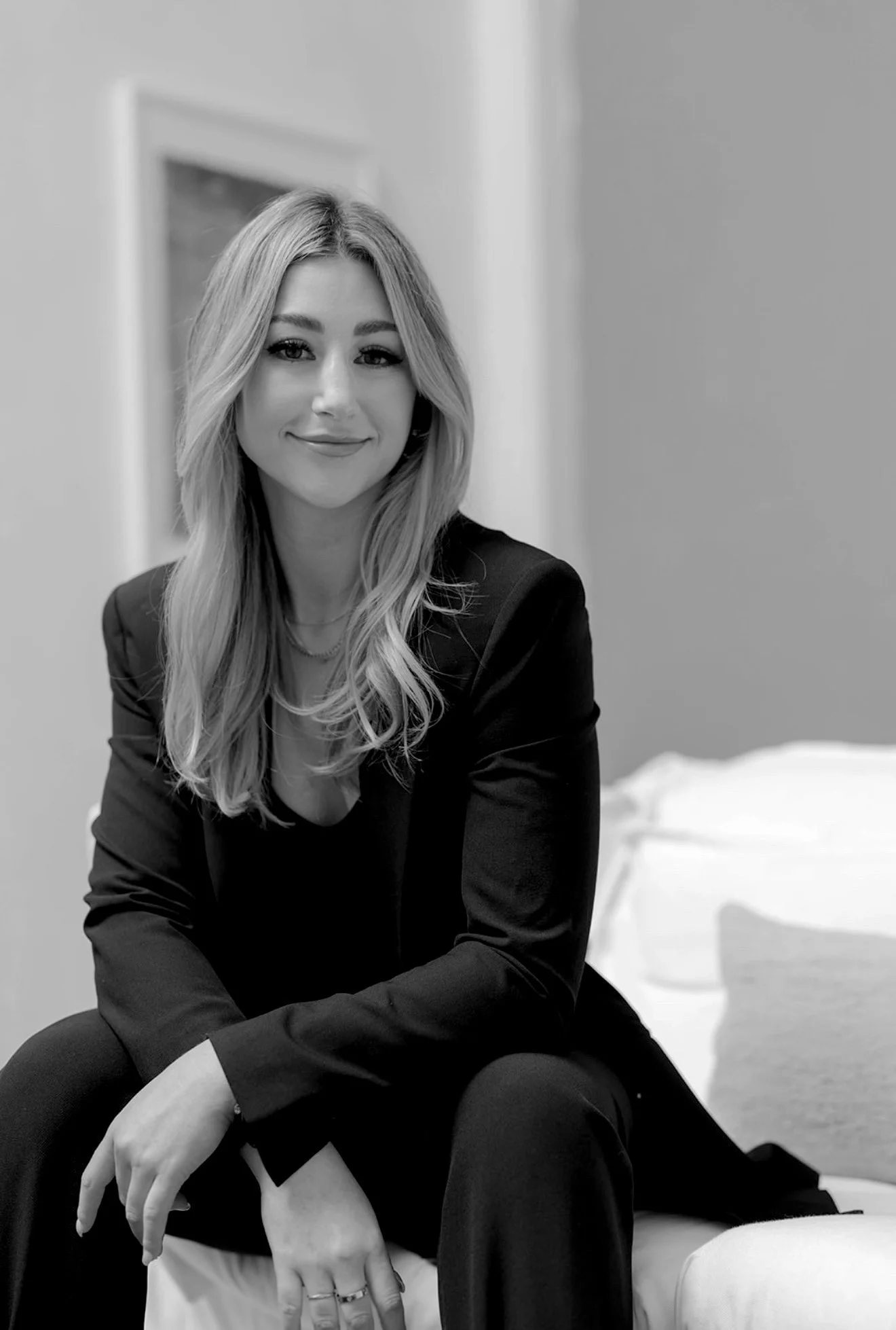 A woman with long, wavy blonde hair, smiling, wearing a black blazer and pants, sitting on a white couch in a room with a blurred background.