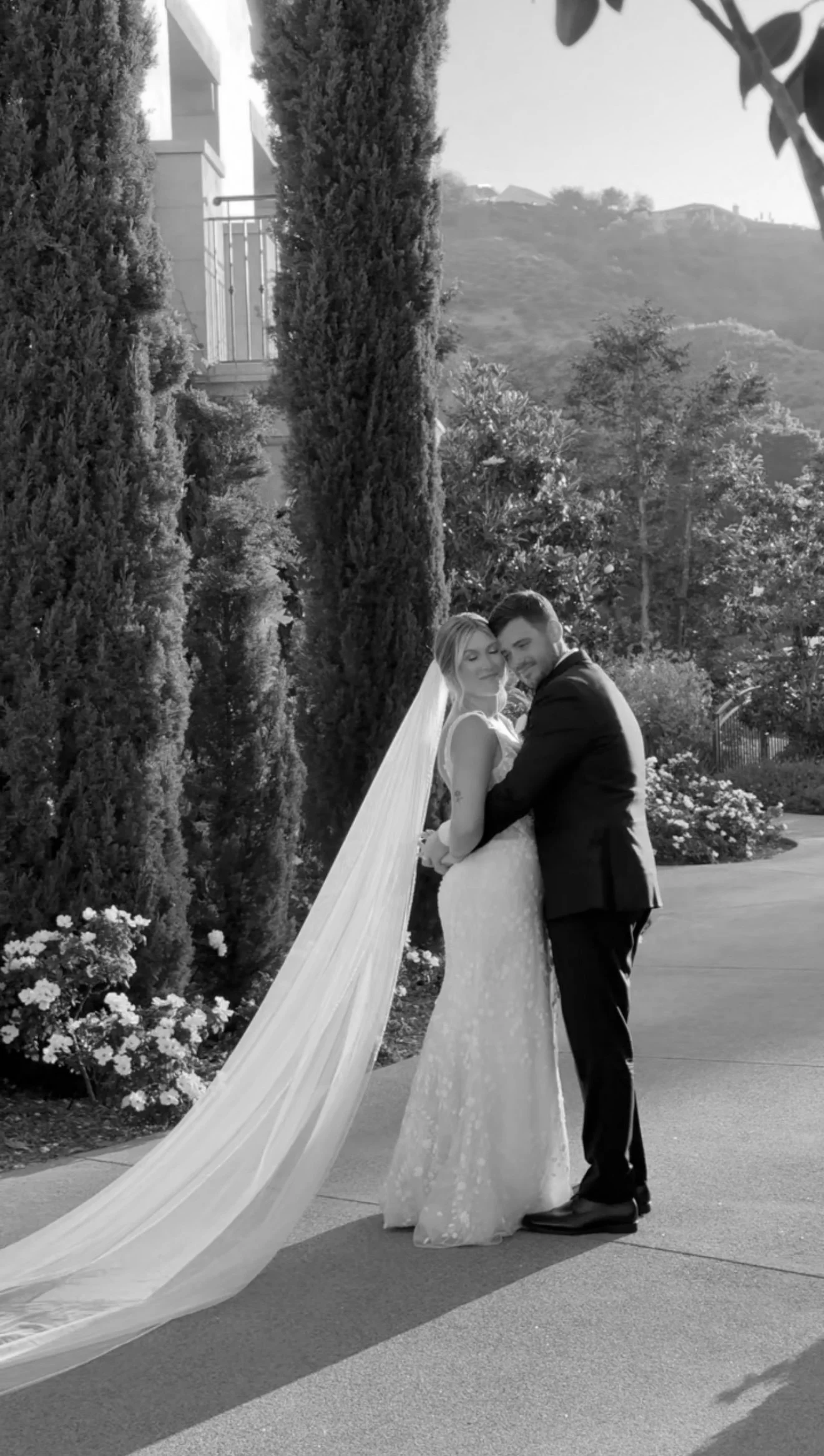 Black and white photograph of a bride and groom hugging outdoors, with decorative trees and mountains in the background.
