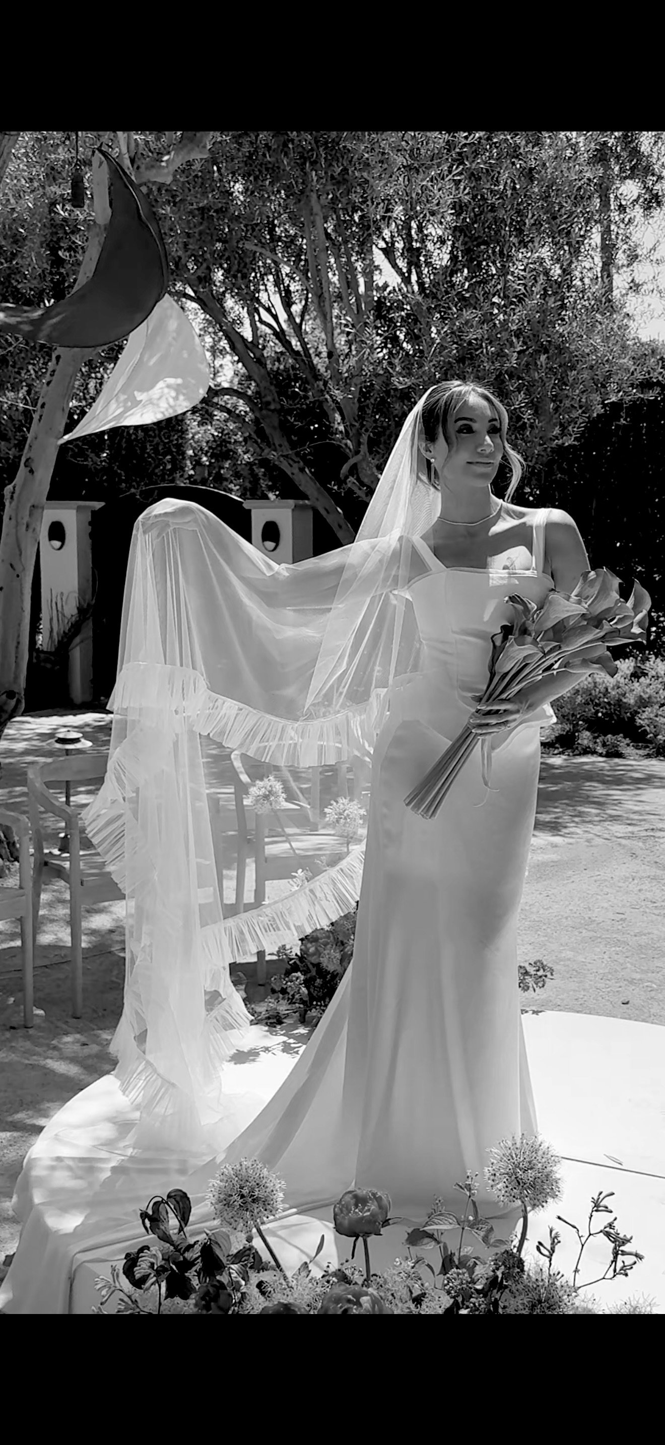 Black and white photo of a bride holding a bouquet of lilies in an outdoor garden setting, with trees in the background and chairs and tables behind her.
