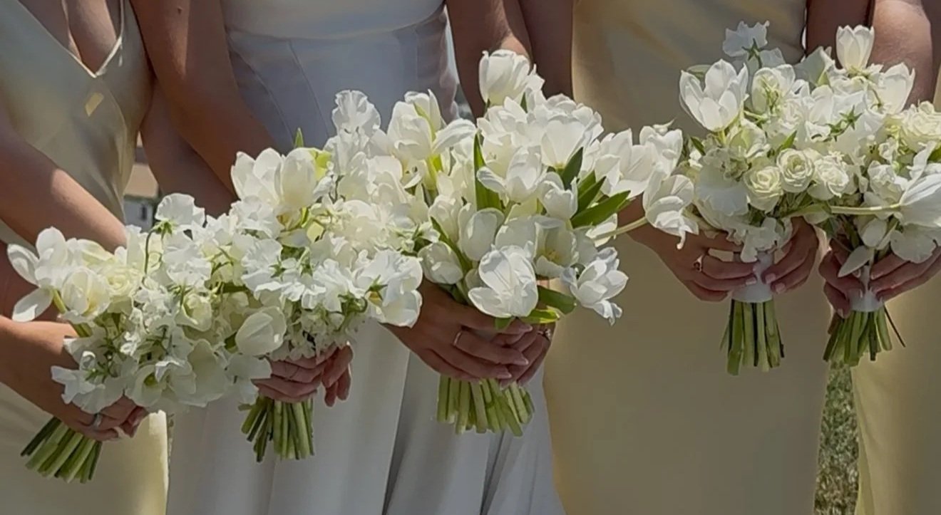 People in pastel-colored dresses holding white floral bouquets at a wedding or special event.