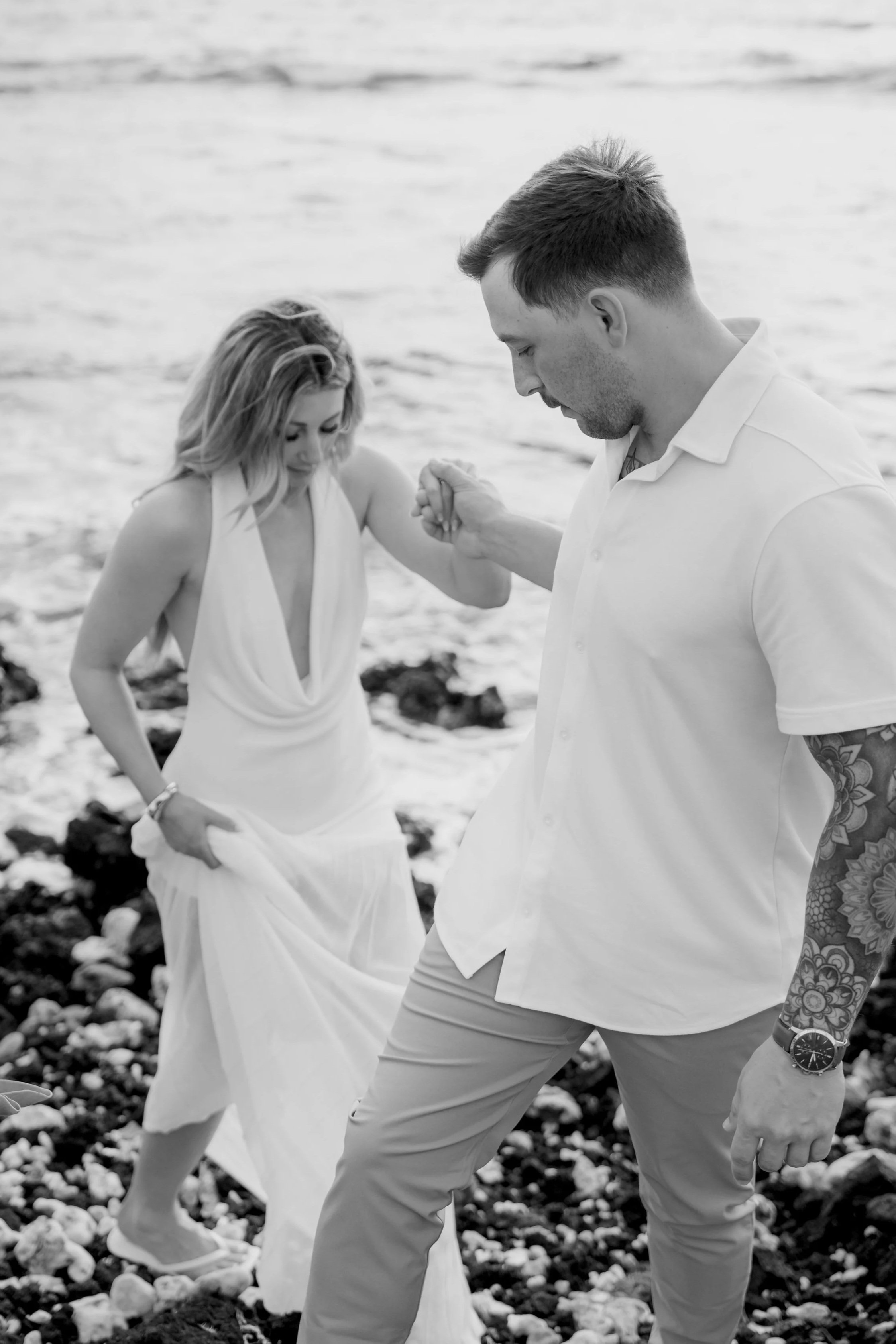 A black-and-white photo of a couple holding hands on a rocky beach, with the ocean in the background. The woman is wearing a white dress and the man is dressed in a light-colored shirt and pants.