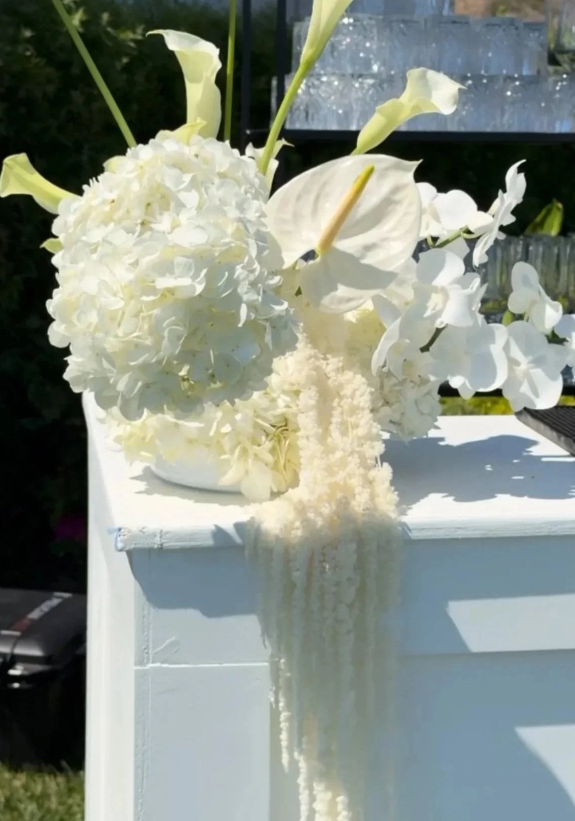 A floral arrangement with white hydrangeas, calla lilies, and anthuriums in a white vase on a white table outdoors.