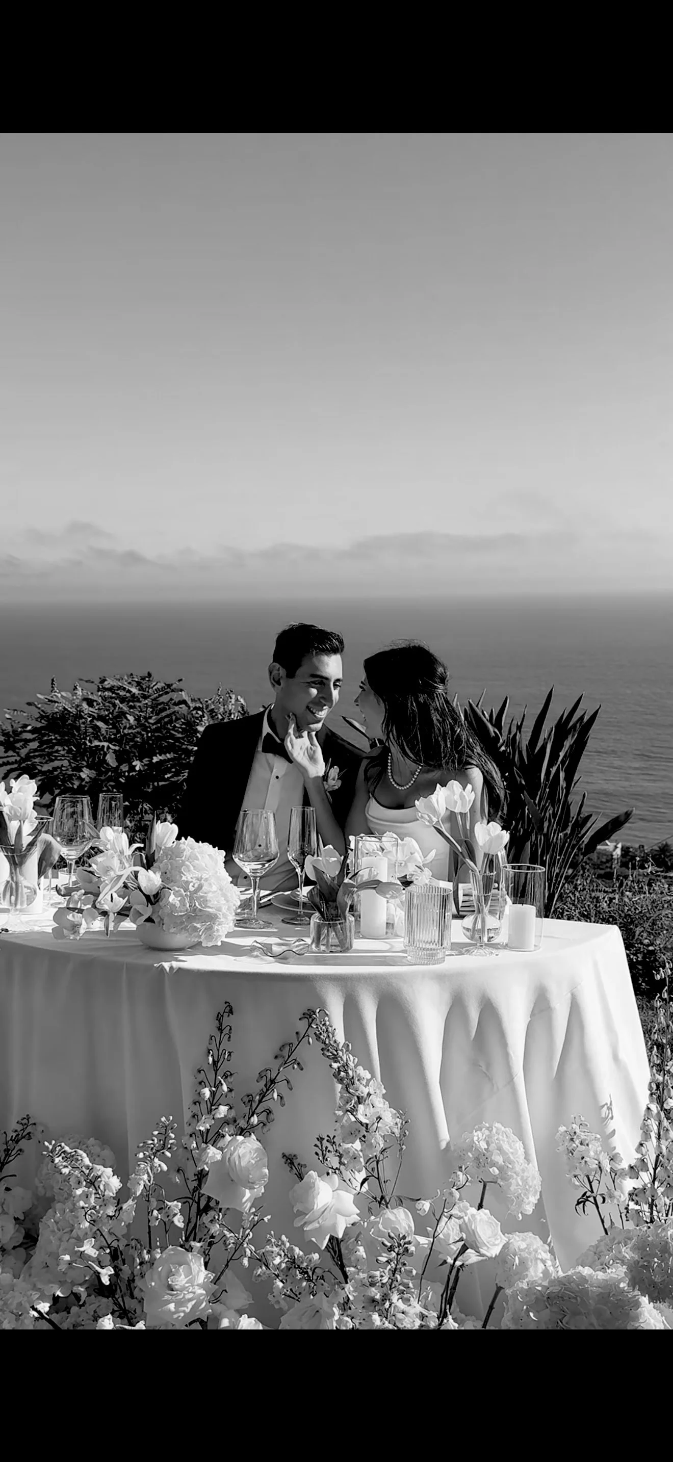 A black and white photo of a newlywed couple at a romantic outdoor wedding reception, sitting at a decorated table with flowers, candles, and glassware, overlooking an ocean view.