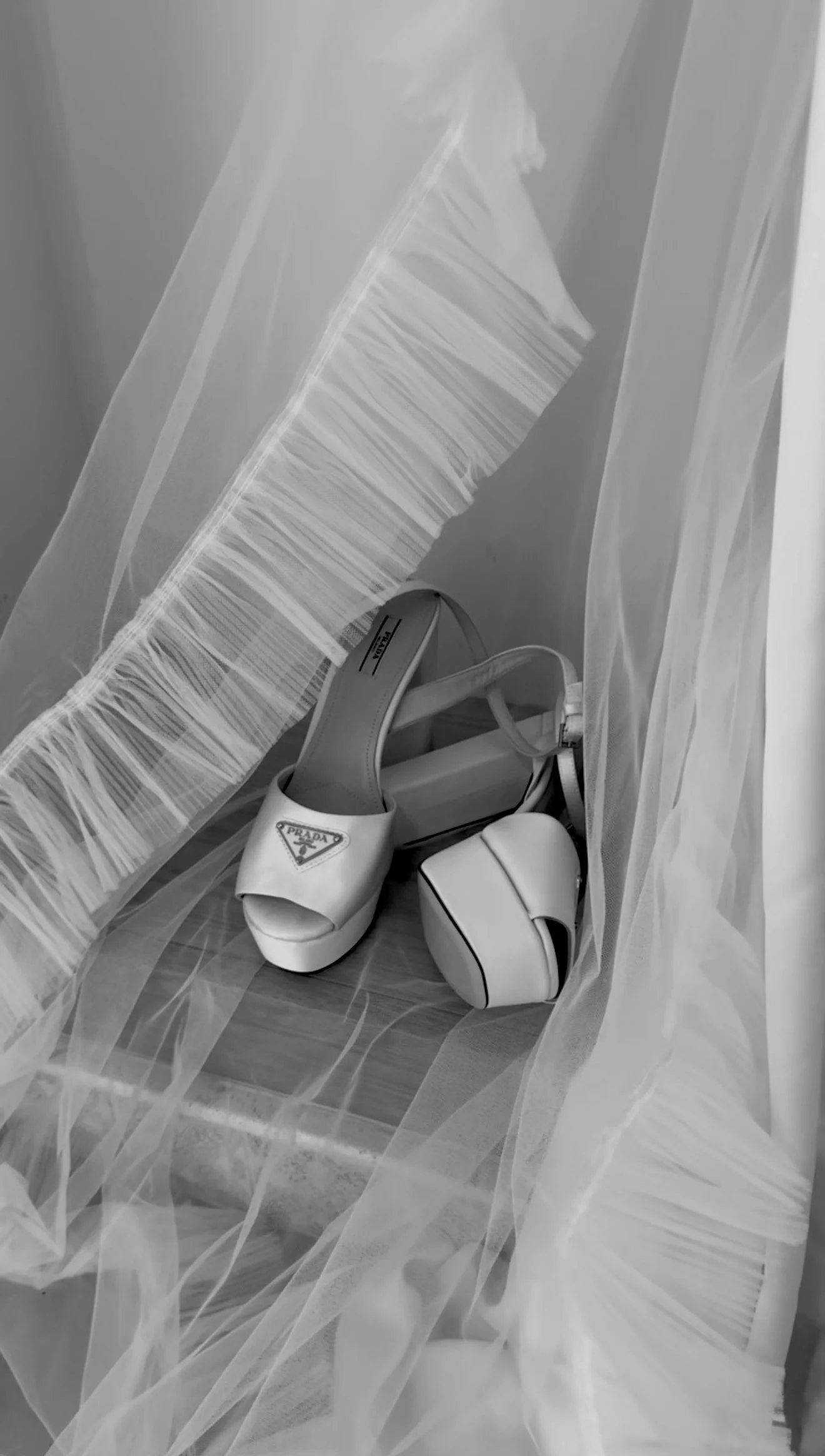 A pair of white Prada high-heeled shoes, a white handbag, and a shoe box inside a transparent plastic bag on a wooden surface.