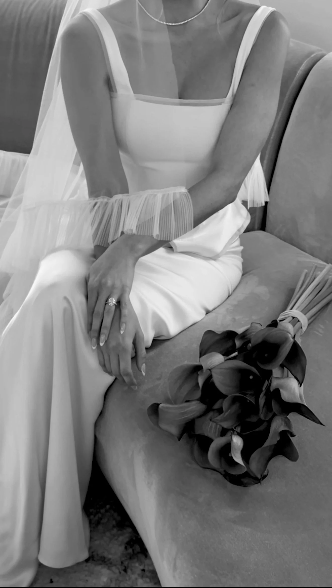 Black and white photo of bride sitting on a couch with a bouquet of flowers beside her. She is wearing a wedding dress with sheer sleeves and a veil, and has a ring on her finger.