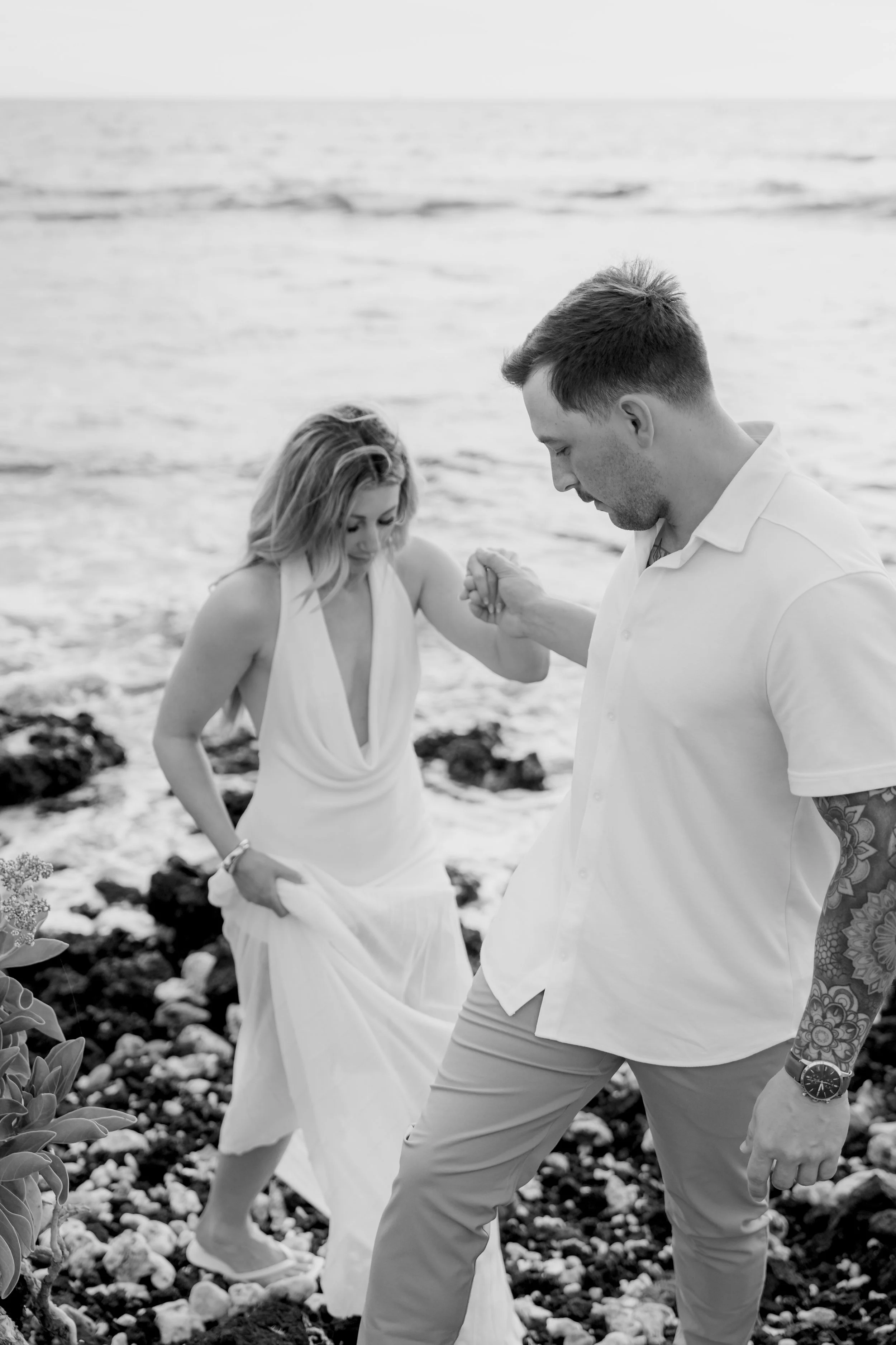 A couple holding hands and walking on a rocky beach, with the ocean in the background.
