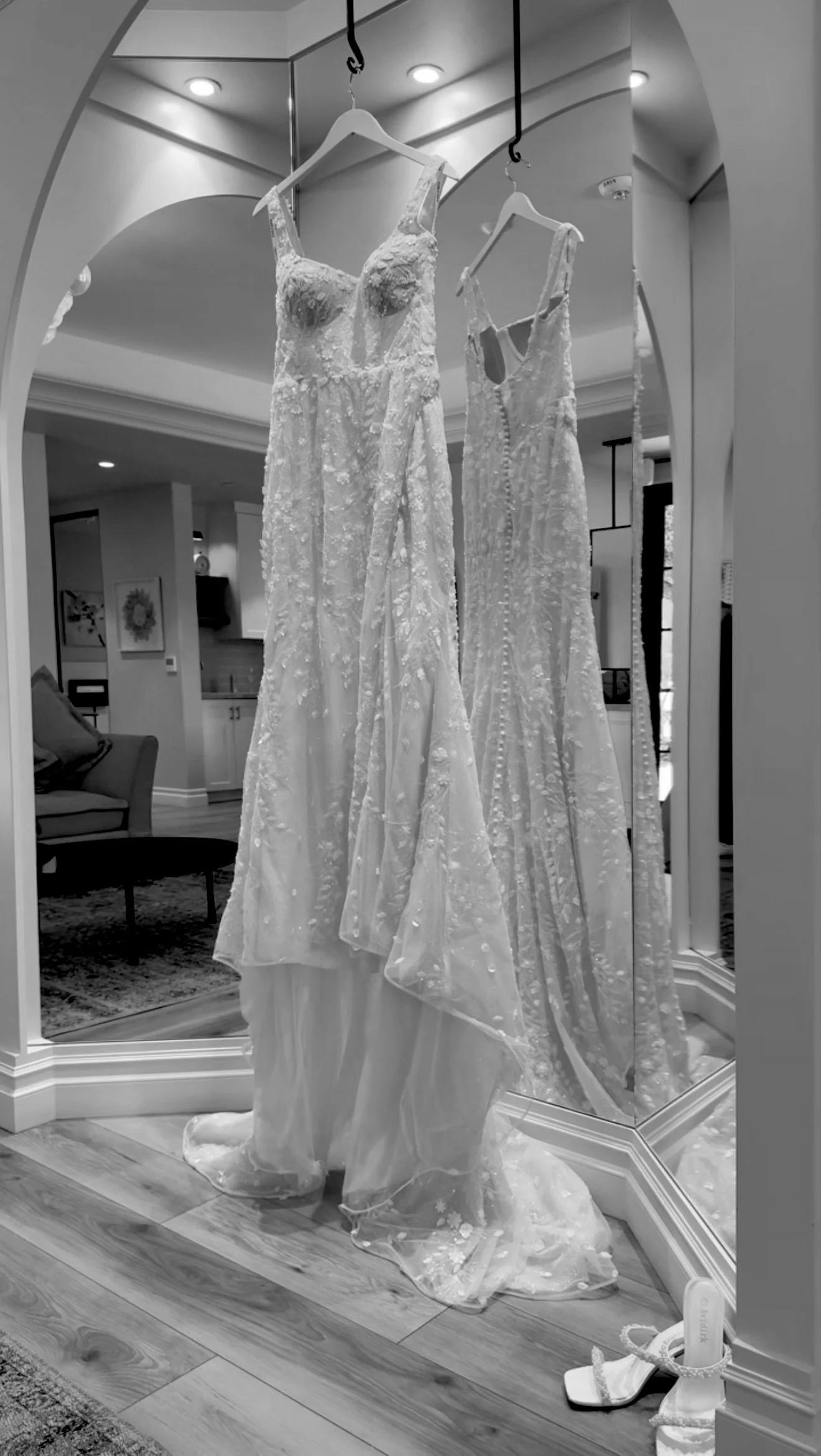 Two wedding dresses hanging on a mirror with a pair of white high-heeled shoes on the floor in front.