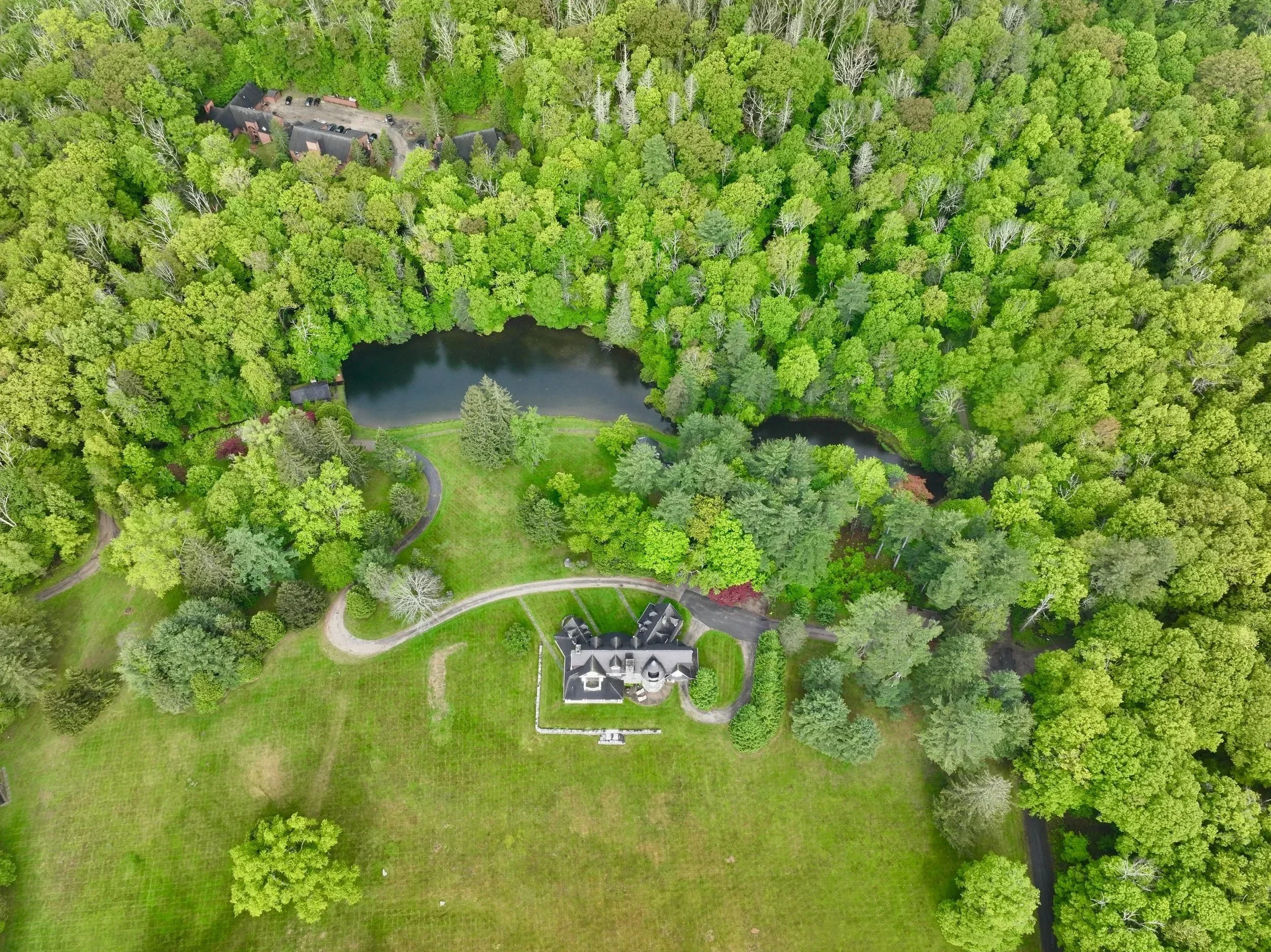 An aerial view of The Thyme Retreat Center with a gray roof, surrounded by lush green trees, a small pond, and winding driveway in a forested area.