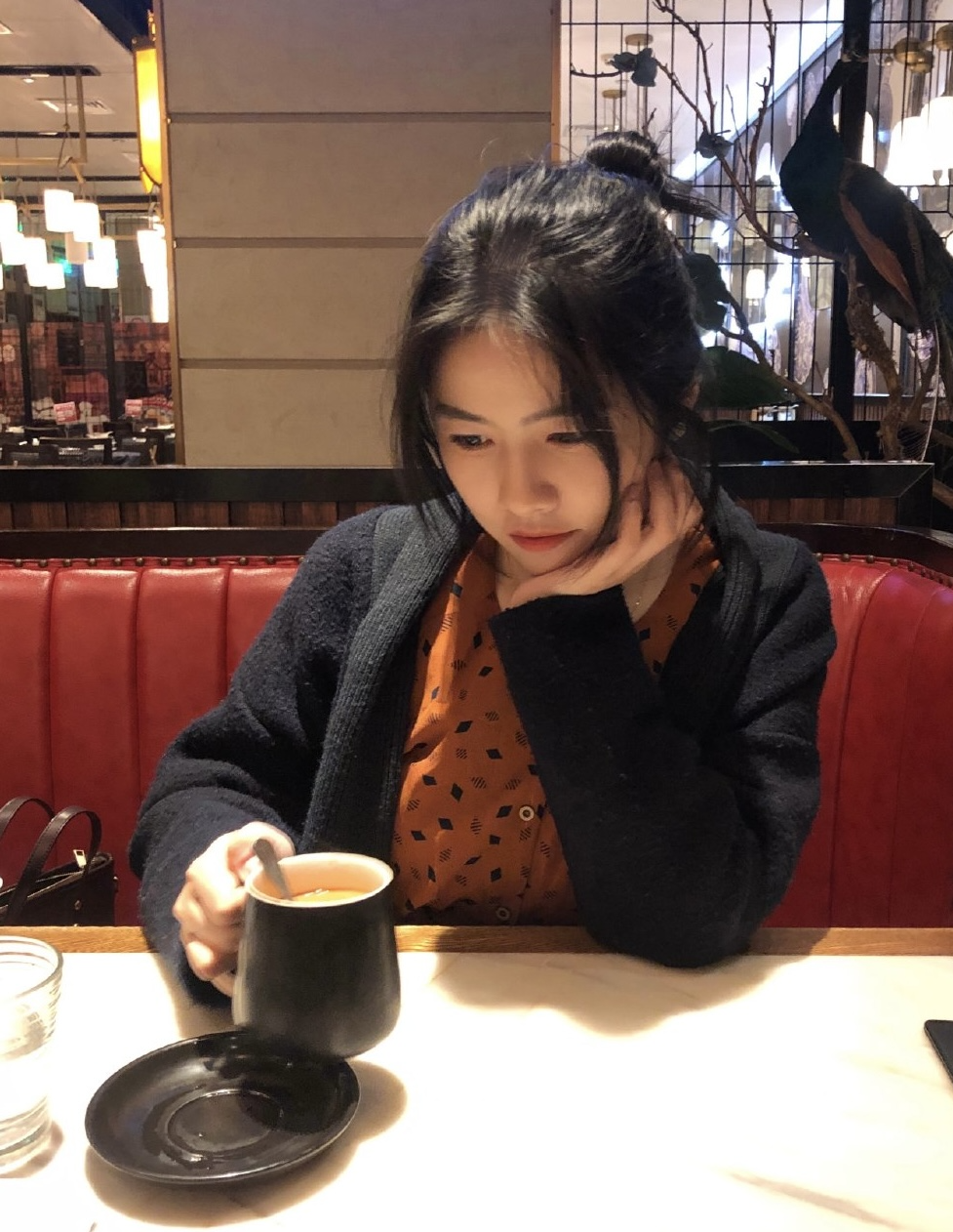 A young woman sitting at a restaurant table, holding a black mug with a spoon inside, and looking at it thoughtfully. She has dark hair tied up, and is wearing a patterned orange blouse and a dark cardigan. The background shows warm lighting, decorative plants, and warm-colored seating.