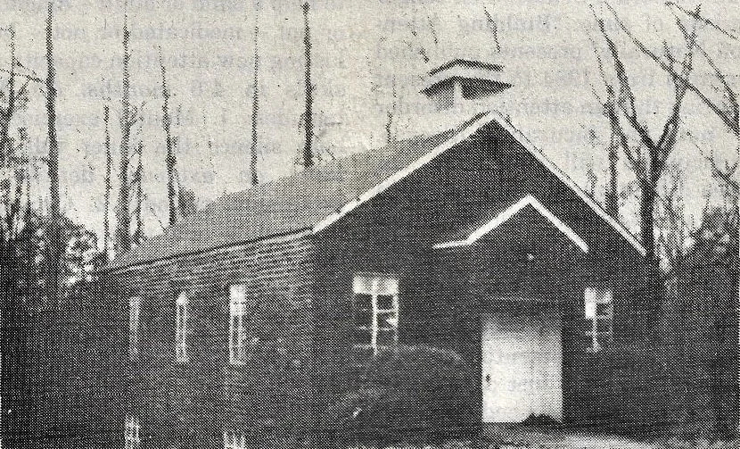 A black and white newspaper photo of a small brick chapel with a steep gabled roof in a wooded area with leafless trees. McKee's Chapel in Dawsonville, GA.
