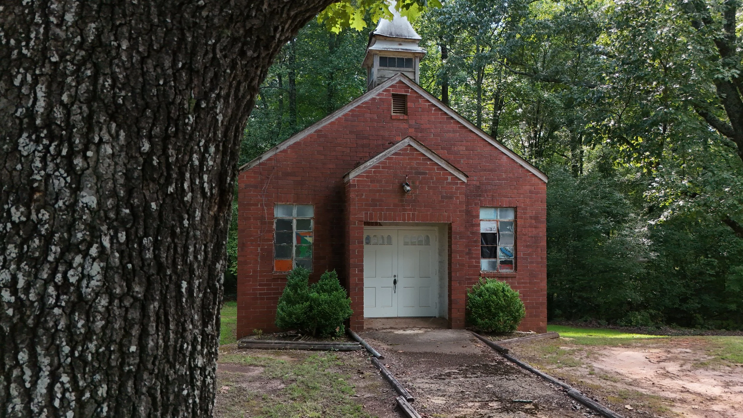 A small brick chapel with white doors, surrounded by trees and greenery, with a dirt path leading to the entrance.