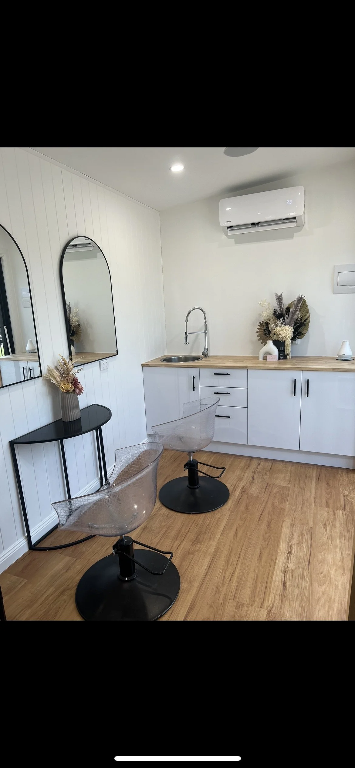 Interior of a modern salon or barbershop with two transparent styling chairs, a black console table with a vase of dried flowers, mirrors on a white paneled wall, a small kitchenette with a sink, decorative flowers, and an air conditioning unit on the wall.