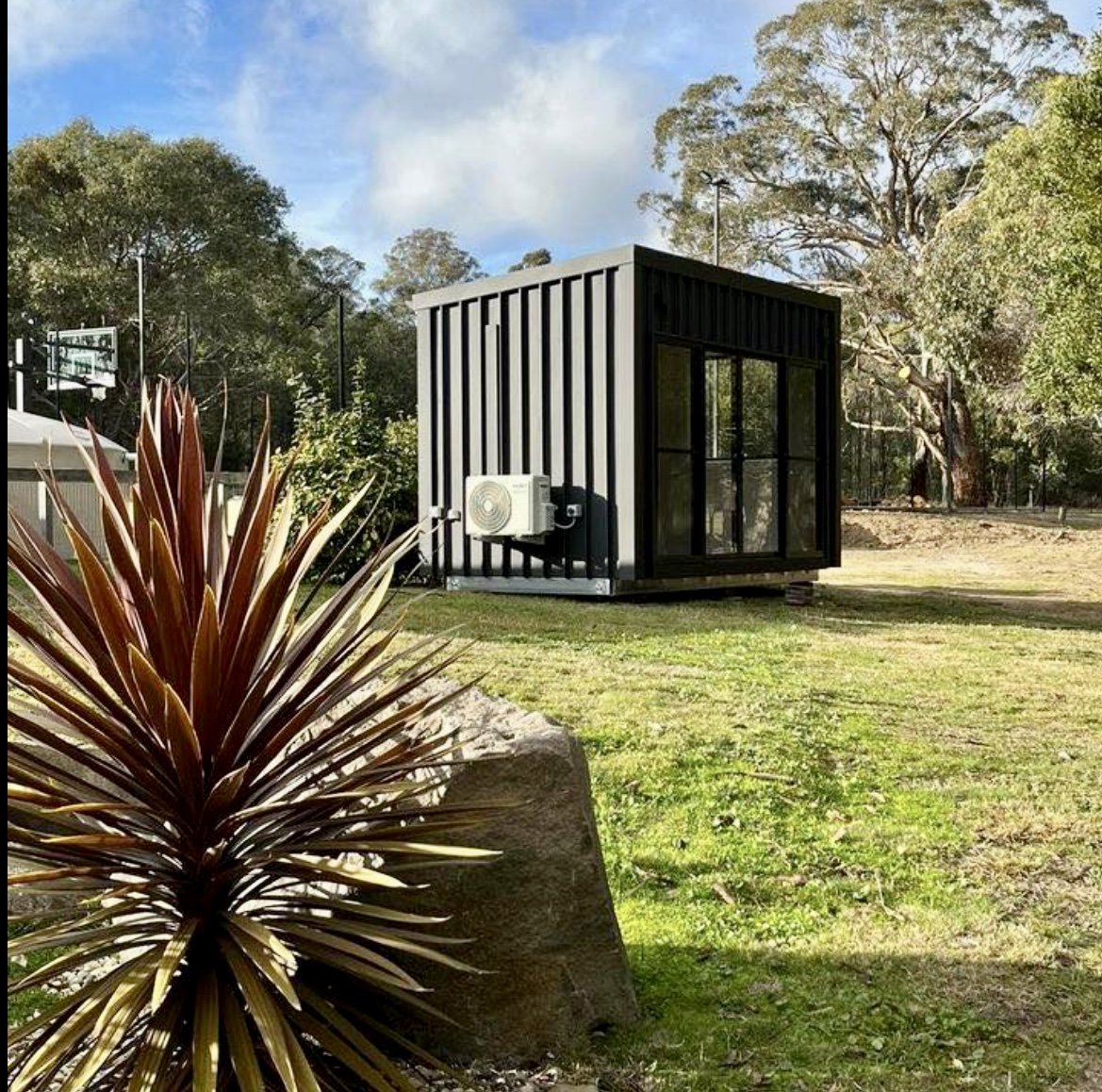 Small modern black container building with glass double doors and an air conditioning unit, situated on a grassy yard with trees and a basketball hoop in the background.