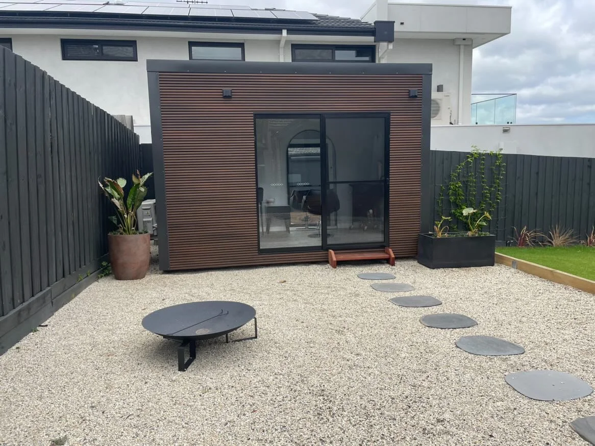 Small modern backyard with gravel ground, stepping stones, a fire pit, potted plants, and a small building with sliding glass doors.