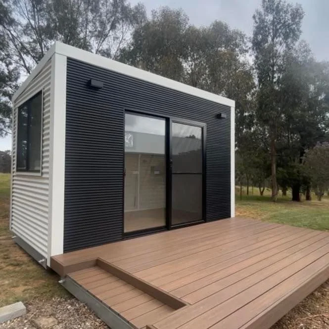 Small modern tiny house with black and white exterior siding and sliding glass doors on a wooden deck, surrounded by trees.