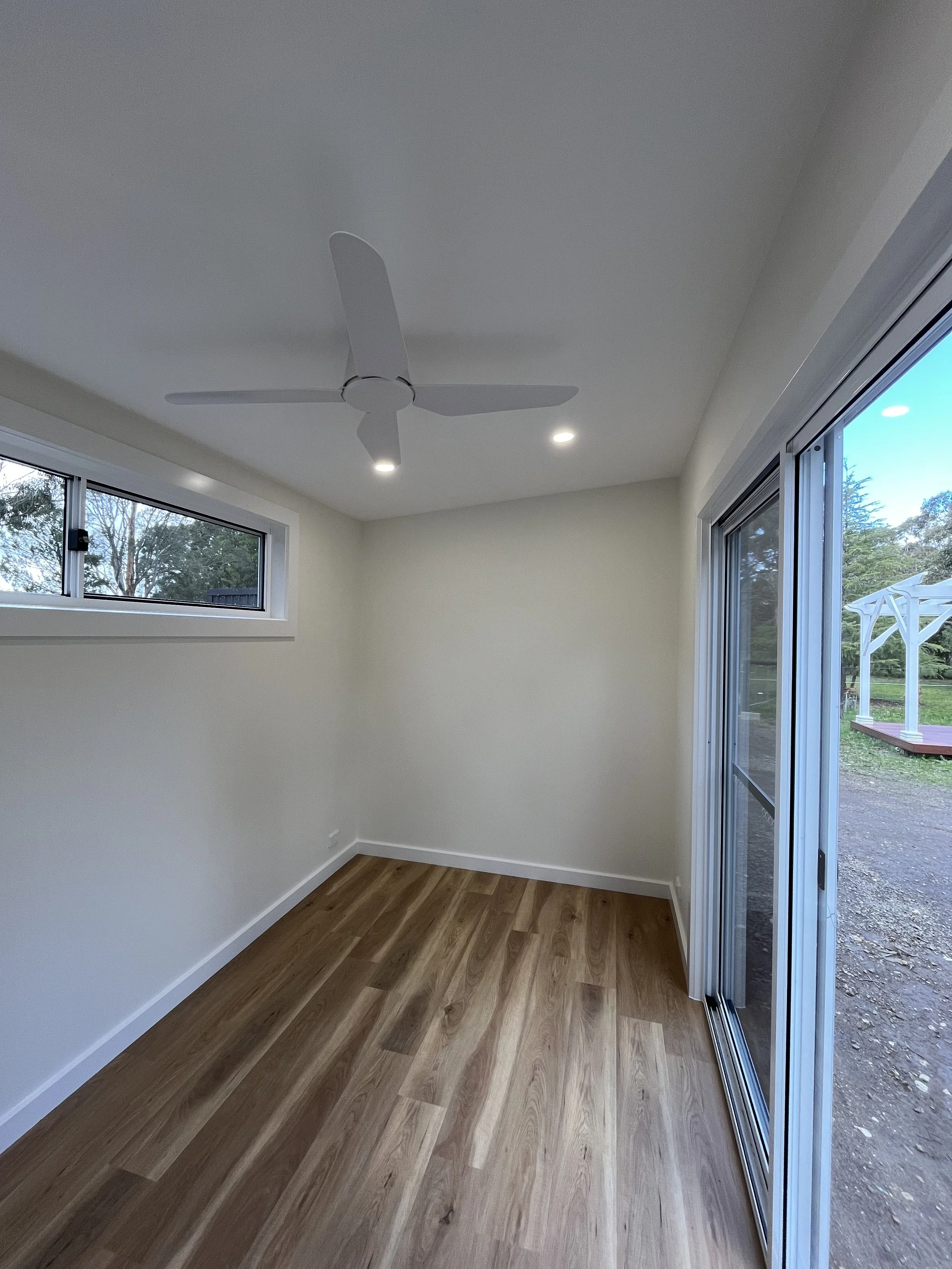 Empty room with white walls, a ceiling fan, small window, sliding glass door leading outside, hardwood floor, and ceiling lights.