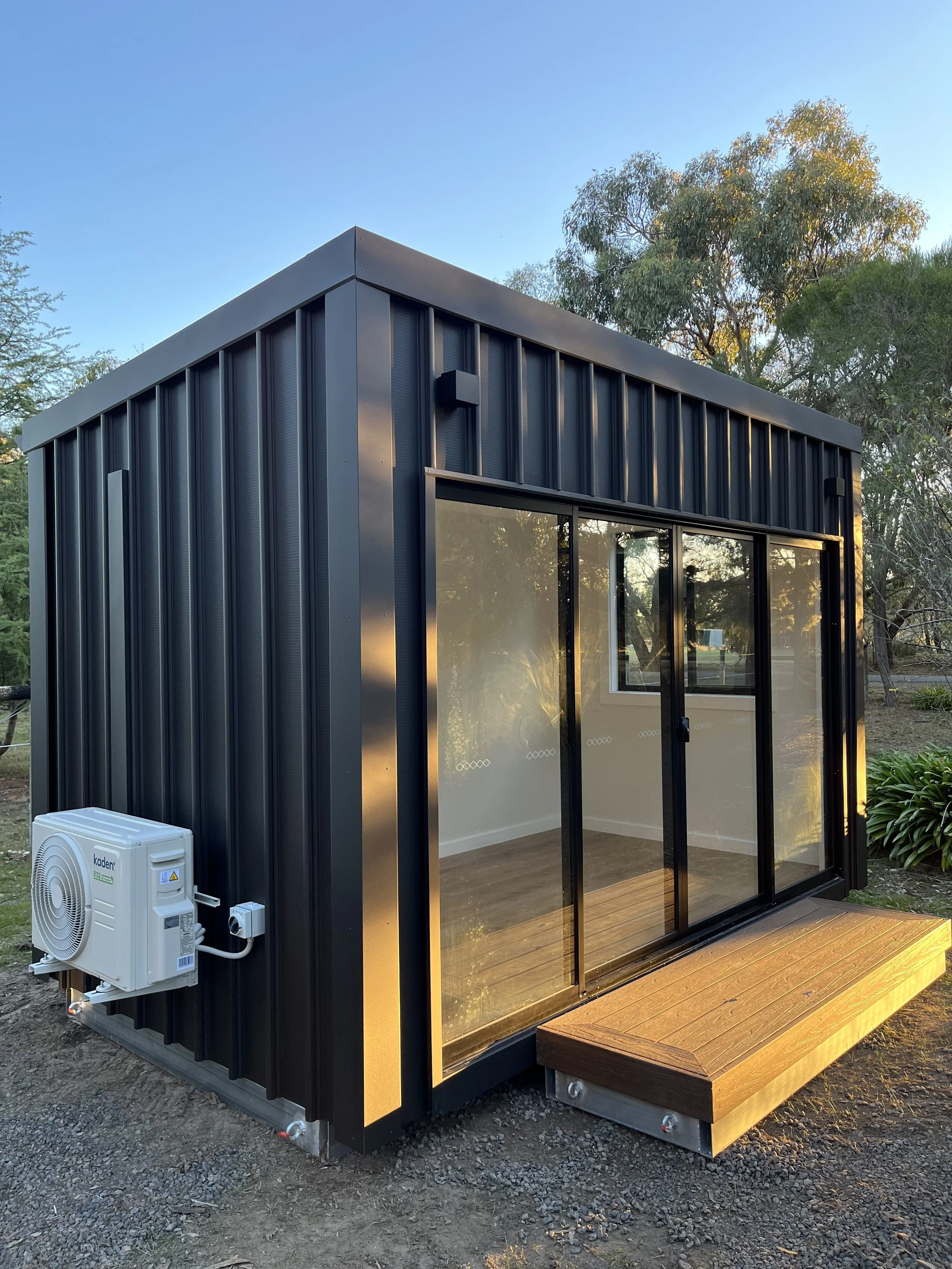 Small modern black metal tiny house with glass sliding doors, wooden deck, surrounded by trees, during sunset