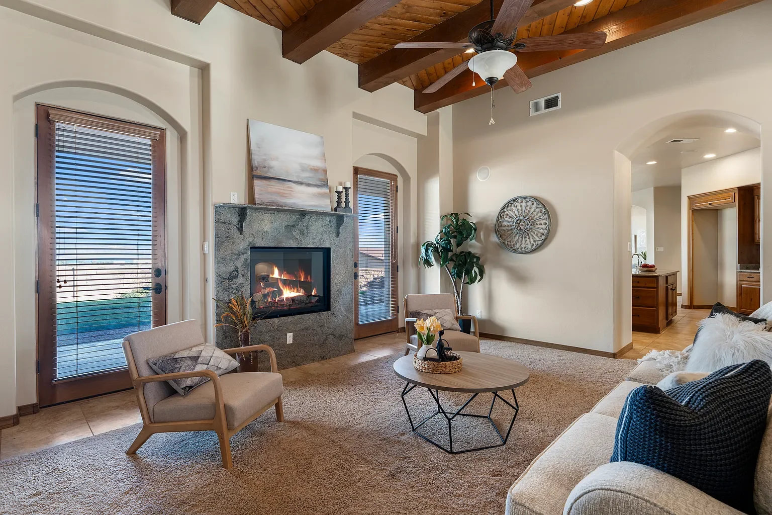 Living room with a wood ceiling, fireplace, glass door, and furniture including a beige sofa, armchairs, a round coffee table, and decorative plants and artwork.