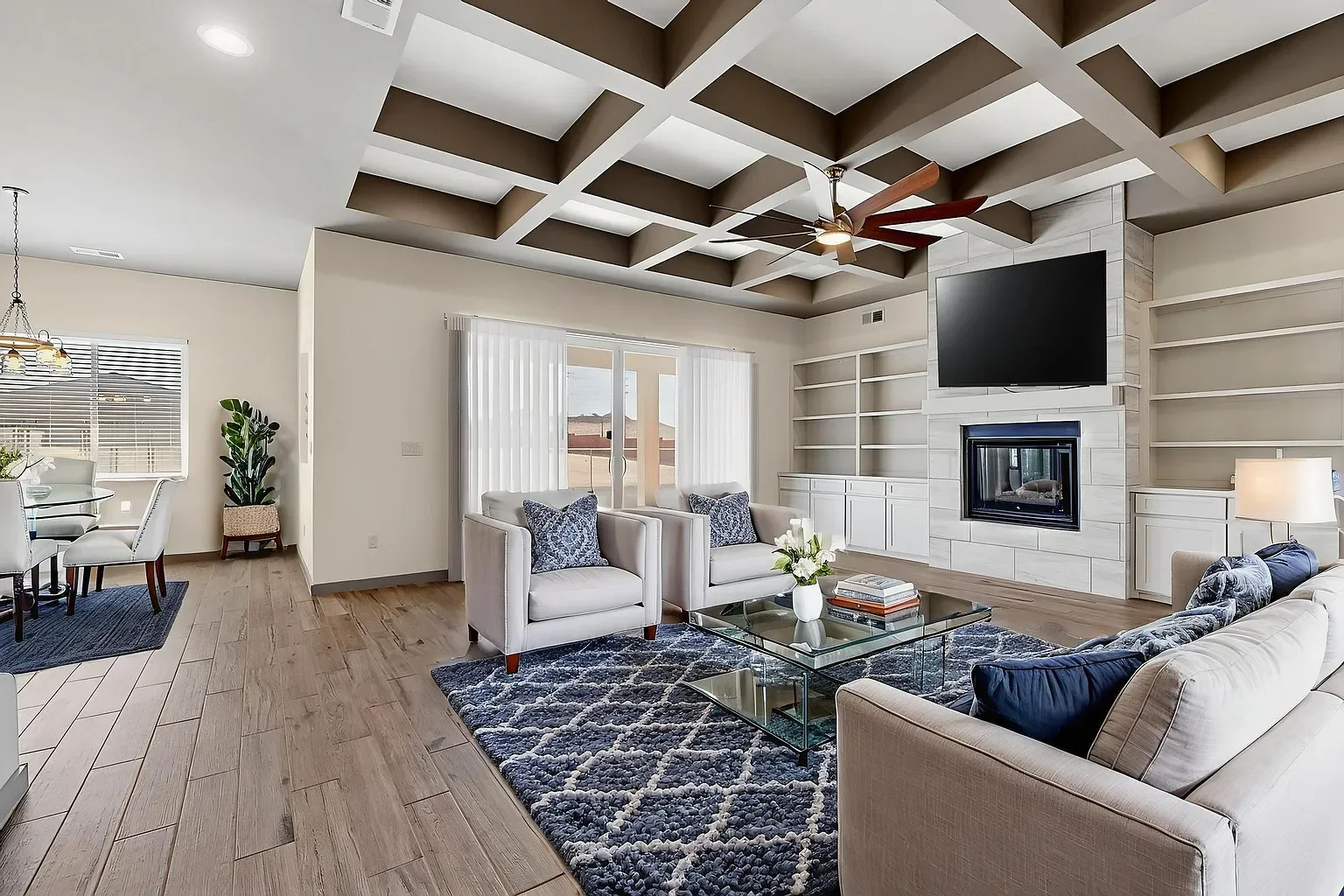 Living room with modern white furniture, a fireplace, and a TV mounted on the wall, beige bookshelves, and a geometric wooden ceiling. There is a patterned blue rug and a glass coffee table with books and a flower vase. Adjacent to the living room is a dining area with a round table and white chairs, a window with blinds, and a potted plant.