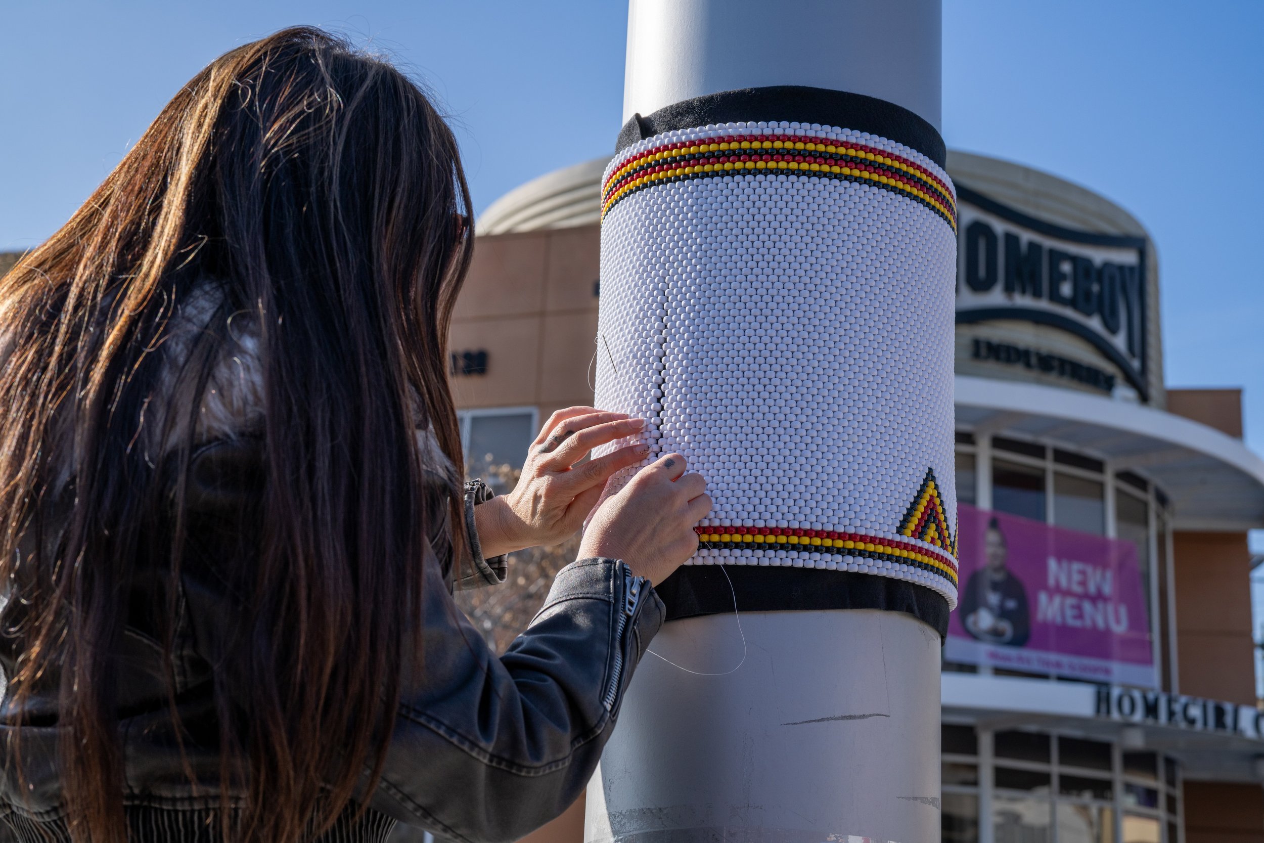 A woman with long brown hair in a black jacket attaches a beaded decoration to a white pole, with a building in the background featuring a sign that reads 'HOME CITY'. The decoration has horizontal yellow, red, and white beads.