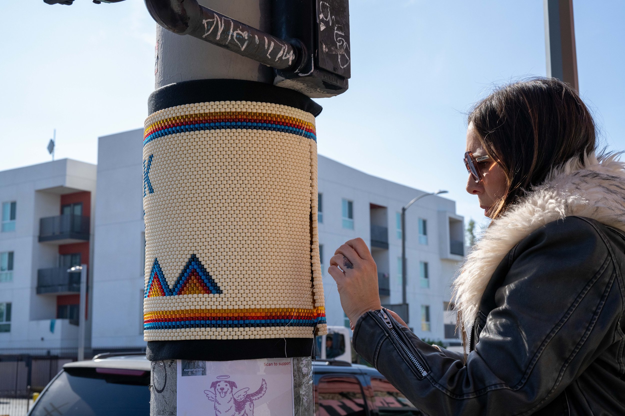 A person with brown hair, sunglasses, and a fluffy fur-lined jacket is attaching or adjusting a beaded decoration on a black pole. The pole has graffiti and a drawing of a dog's face with wings on a piece of paper below it, attached to a sign.