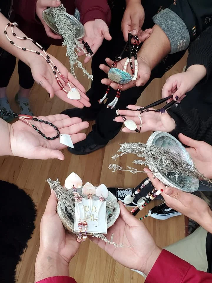 Several hands holding necklaces, shells, and small decorative items, with a wooden floor in the background.