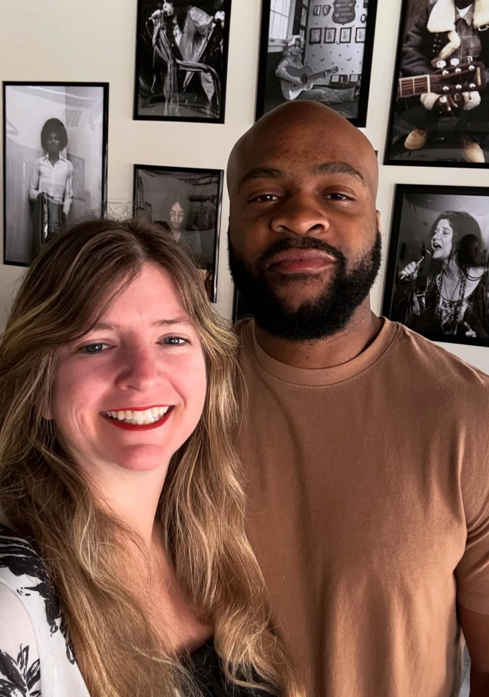 A smiling woman with long, wavy blonde hair and a man with a beard and bald head smiling indoors in front of a wall of black-and-white framed photographs, including images of musicians and children.