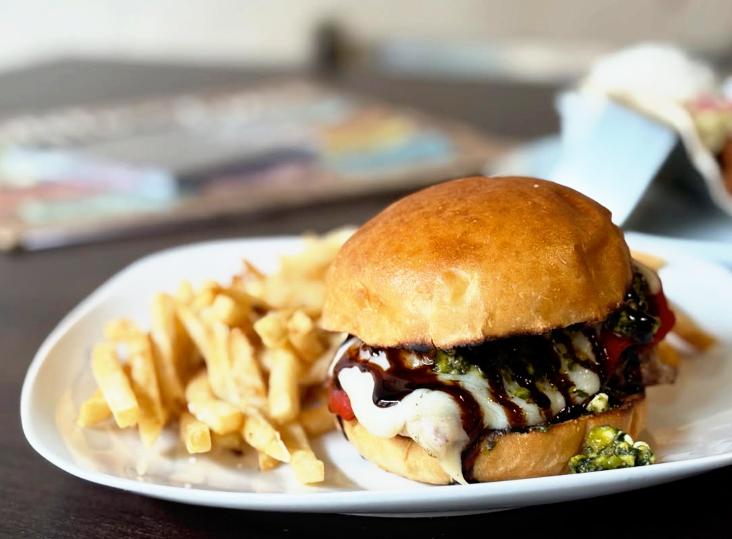 A sandwich with a toasted bun, filled with cheese, vegetables, and a dark sauce, served with a side of French fries on a white plate.