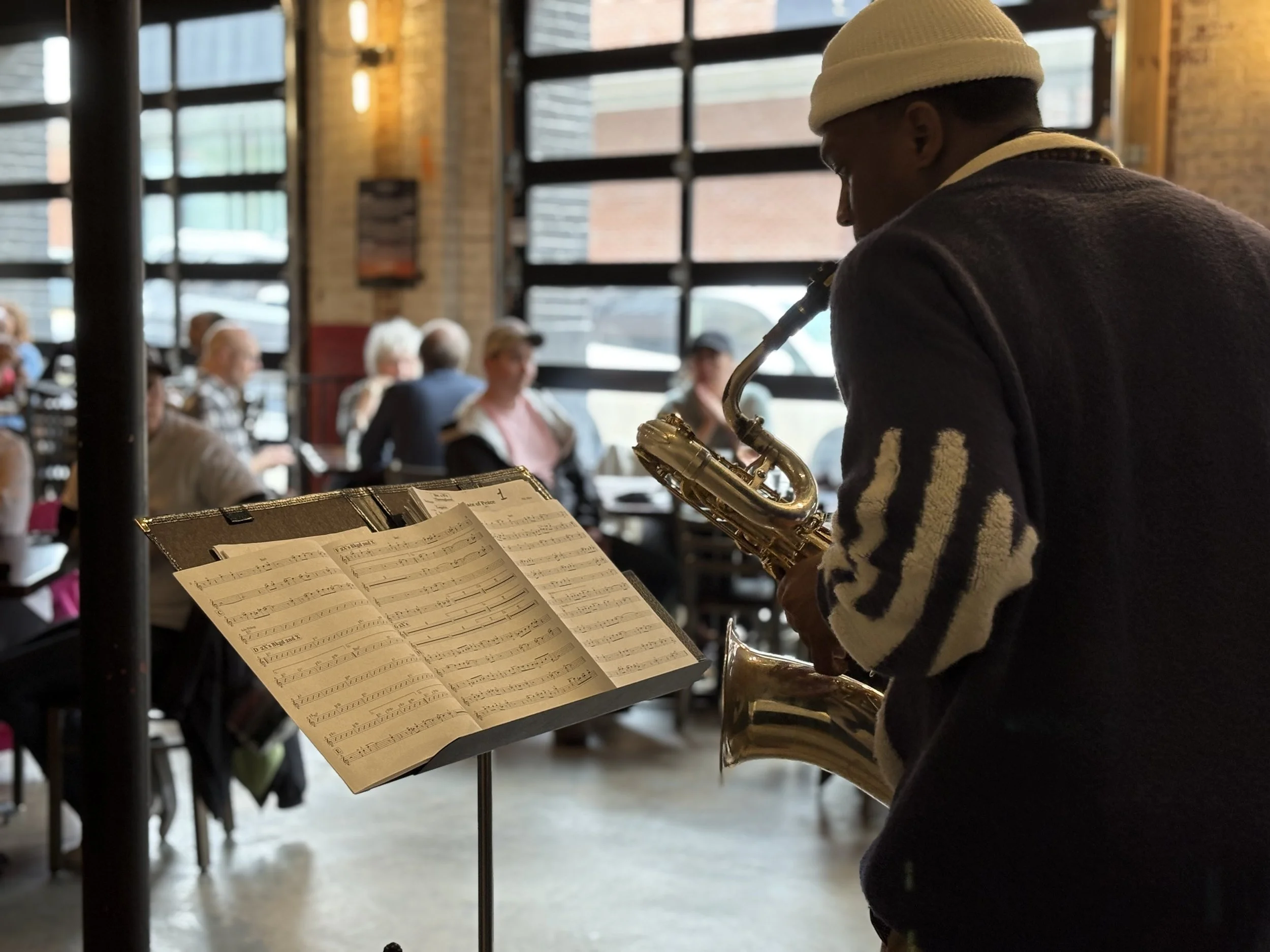 A person dressed in a dark sweater and white beanie playing a saxophone in a cafe or restaurant. The sheet music is visible on a stand in front of them. The background shows several people sitting at tables by large window panes, with warm lighting and a brick wall.
