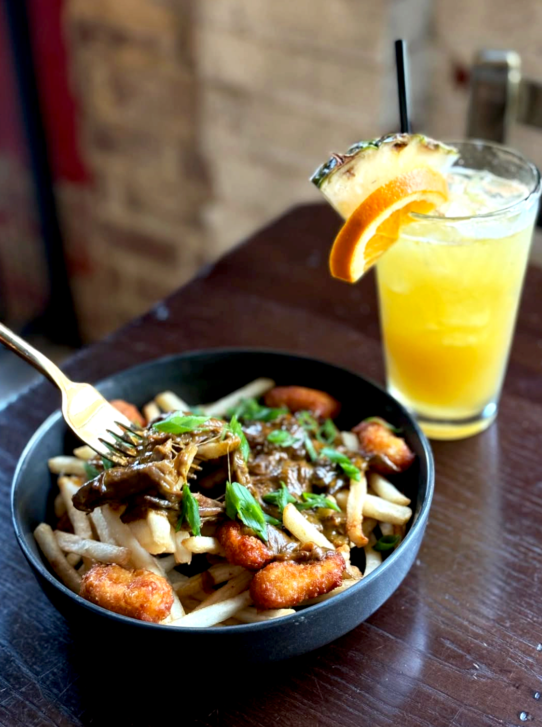 A bowl of pasta with chicken, green onions, and sauce, accompanied by a glass of orange juice with a slice of orange and pineapple.