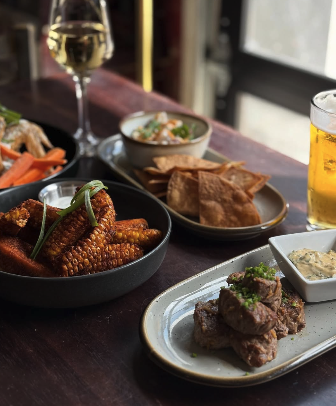 A variety of plated dishes on a wooden table, including corn ribs, salads, chips with dip, and meat with sauce, with drinks of wine and beer in the background.