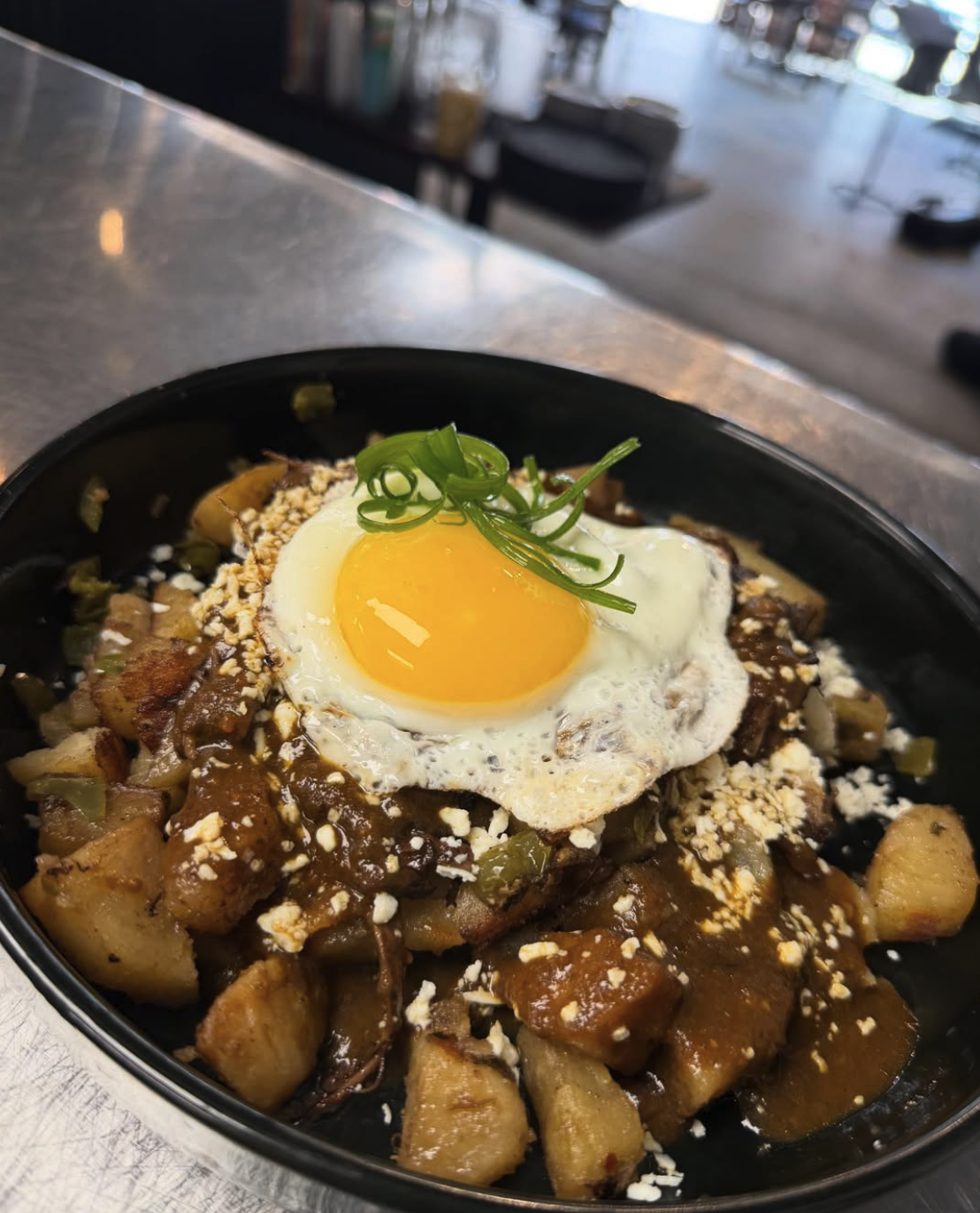 A bowl of food with diced potatoes, shredded meat, brown sauce, crumbled cheese, a fried egg with a runny yolk, and chopped green onions on top.