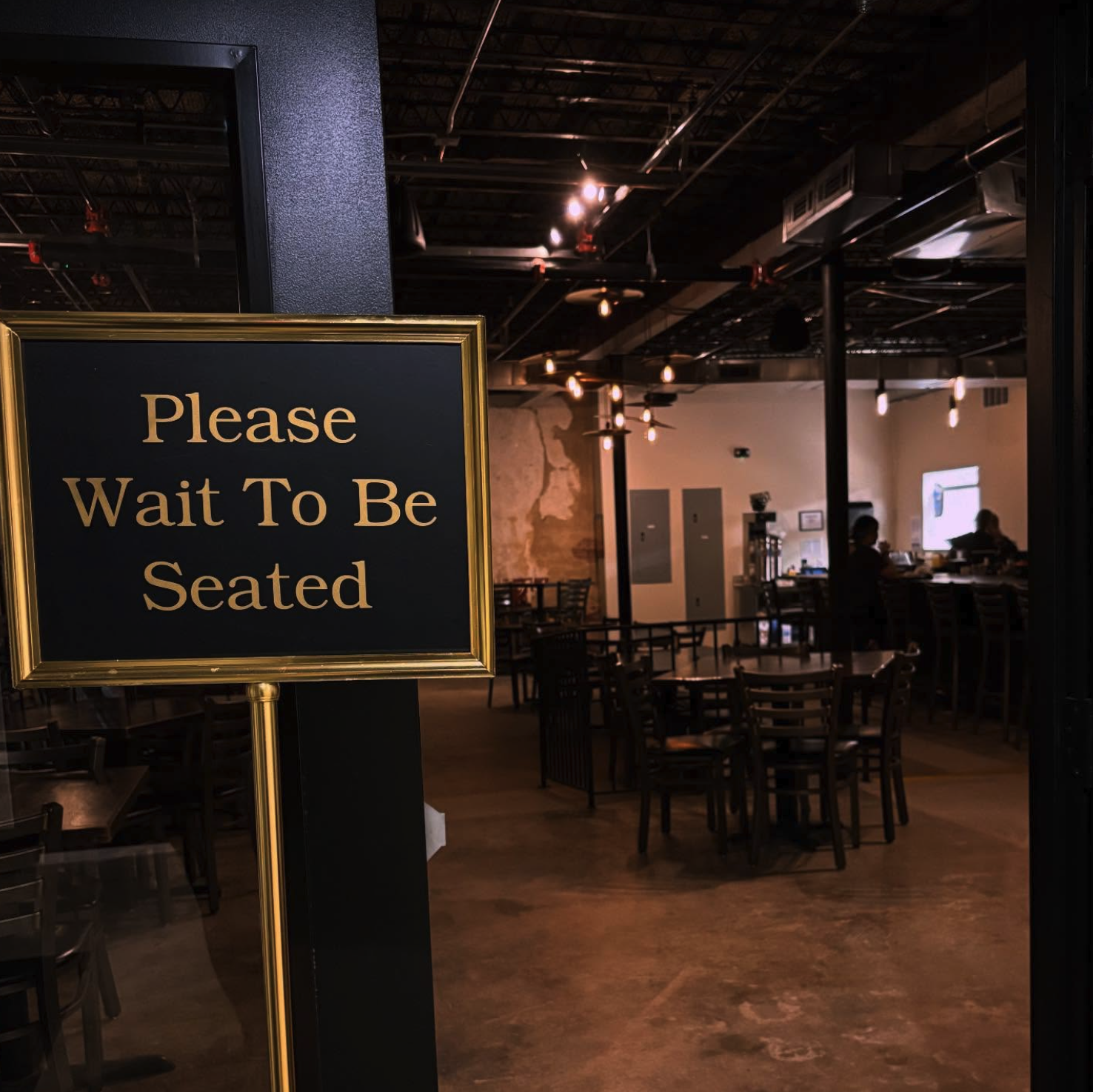 Indoor restaurant scene with a sign that reads 'Please Wait To Be Seated' in the foreground, several empty tables and chairs, and a few people visible in the background.
