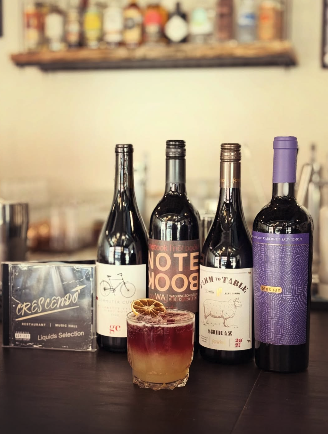 A cocktail with a slice of dried orange, four bottles of wine and a CD on a dark bar counter.