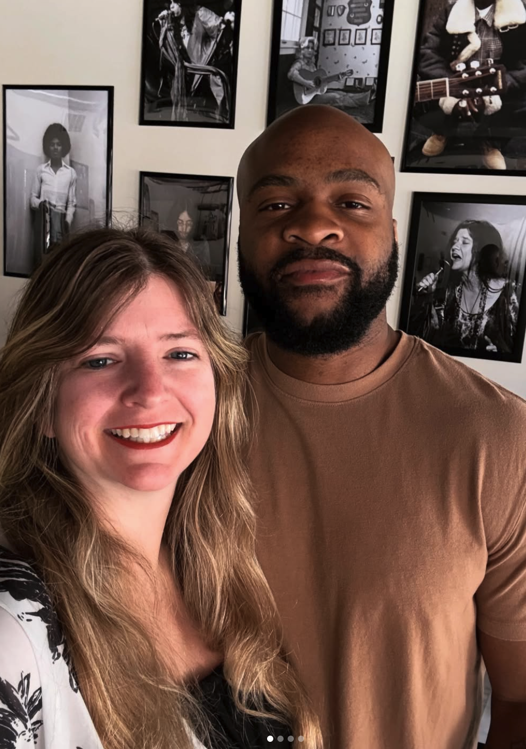 A smiling woman with long blonde hair and a man with a beard taking a selfie together in front of a wall decorated with black and white photographs of musicians and people.