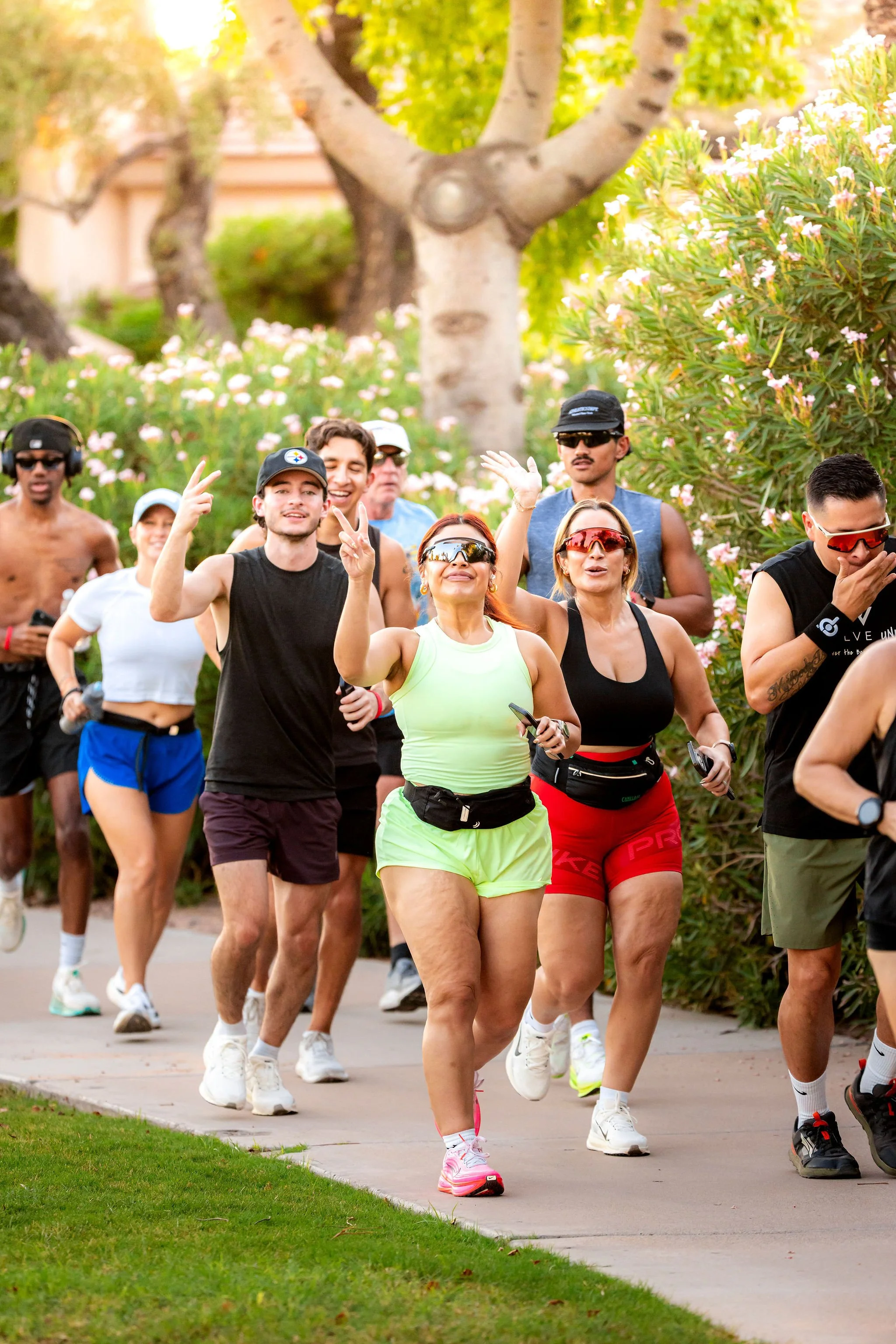 A group of diverse people jogging outdoors on a sunny day, surrounded by trees and flowering bushes.