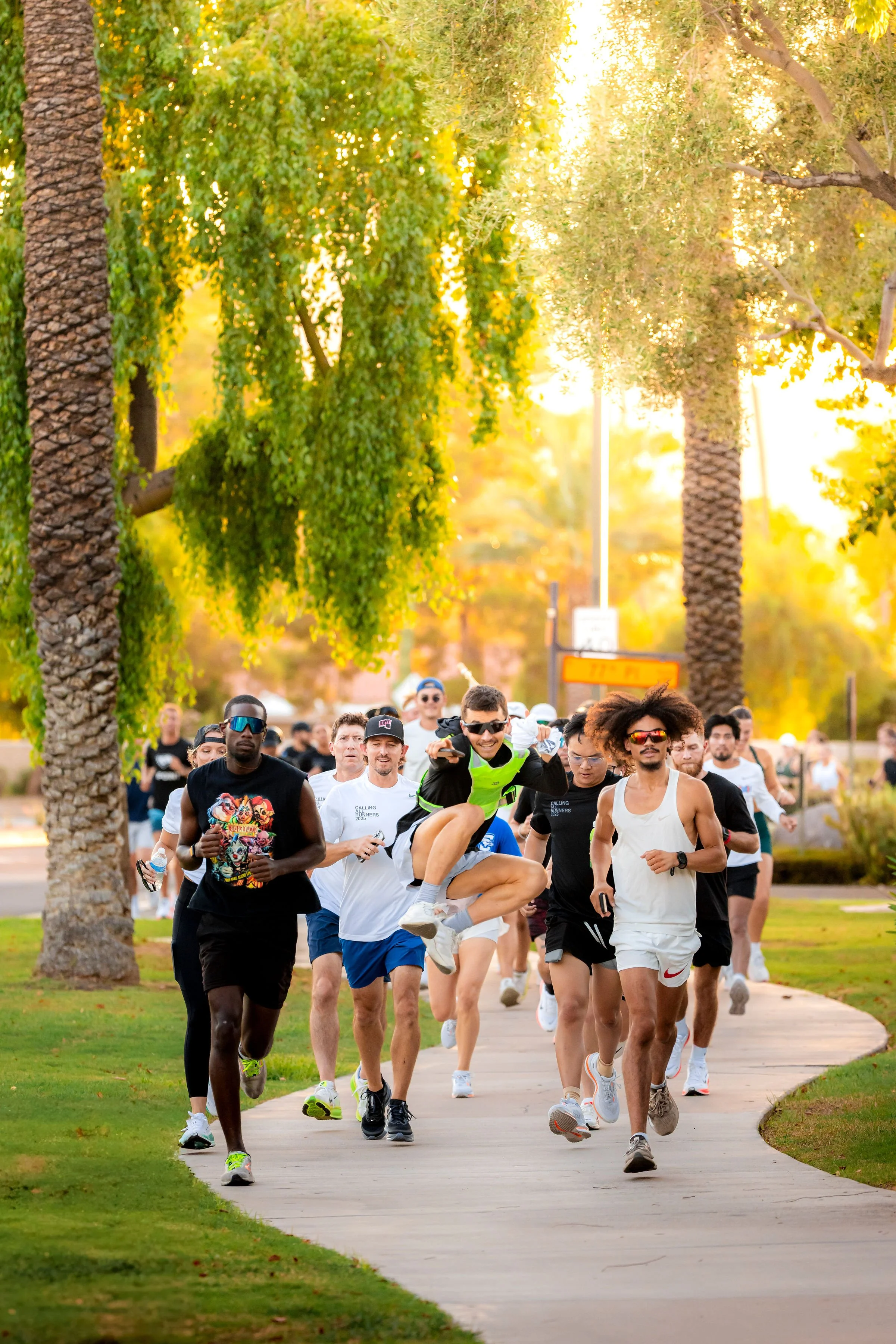 A group of people jogging and running together on a curved sidewalk in a park surrounded by tall trees and greenery during sunset.