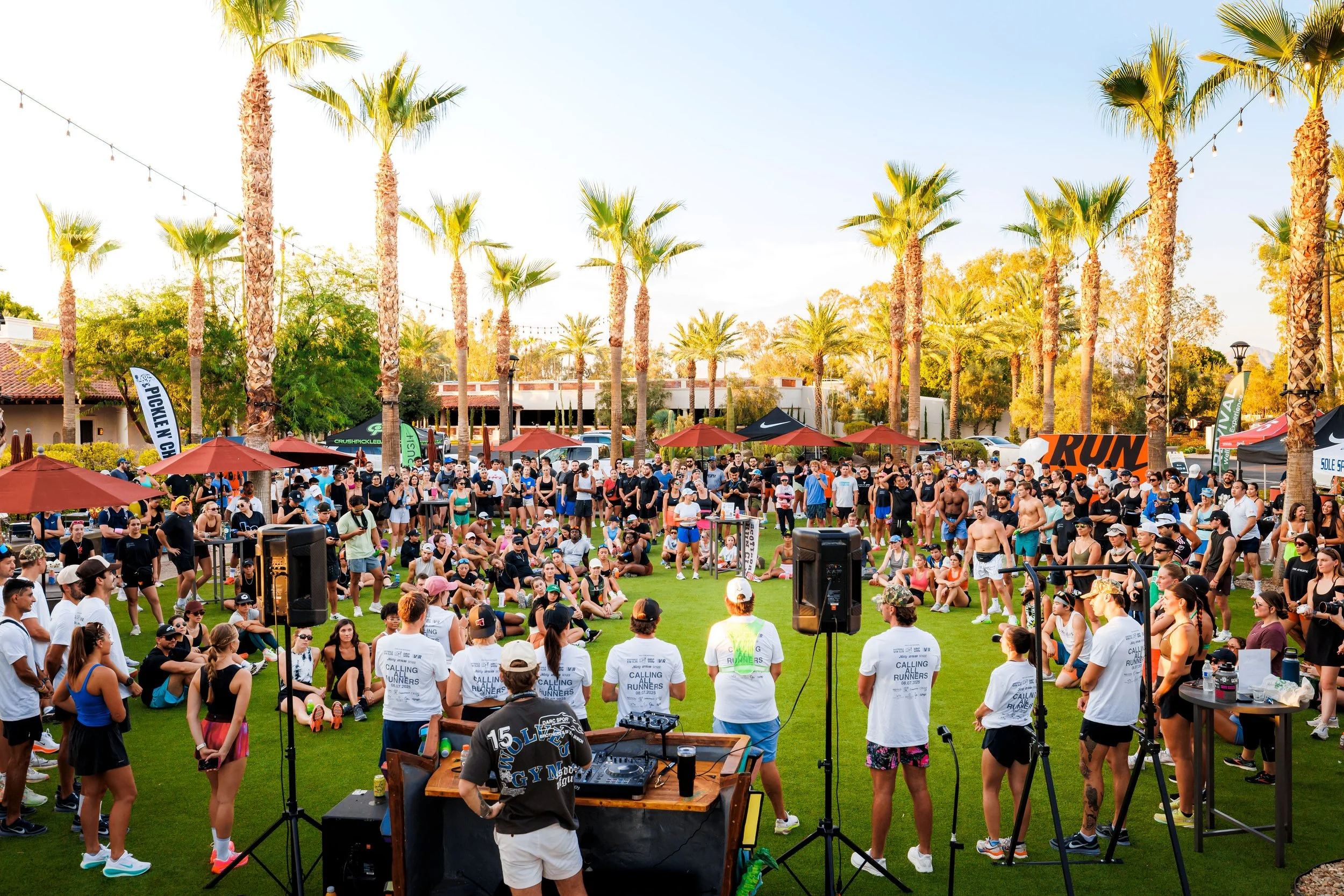 Large outdoor gathering in a park with palm trees, people sitting and standing, some wearing athletic clothing, event banners, and a DJ setup in the foreground.