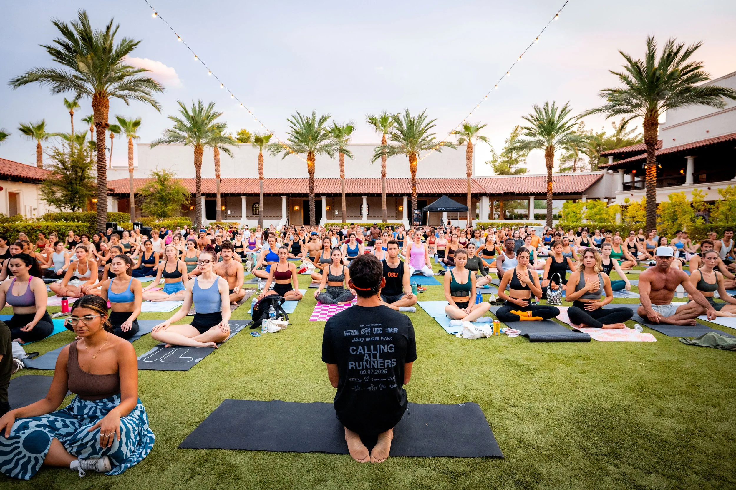 Group of people participating in an outdoor yoga class on a grassy lawn with palm trees and string lights during sunset.